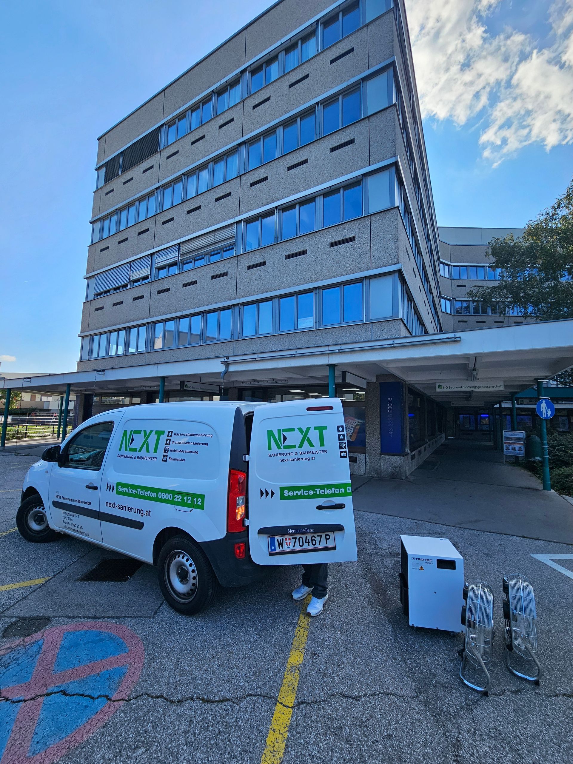 Ein weißer Lieferwagen mit der Aufschrift „NEXT“ parkte vor einem mehrstöckigen Backsteinbürogebäude, auf dessen Boden Ausrüstung stand.