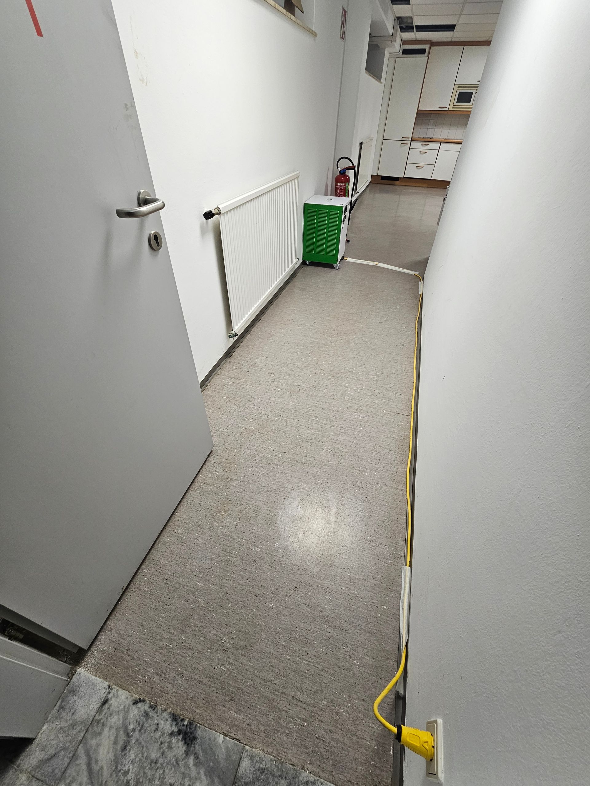 Ein schmaler, schwach beleuchteter Flur mit weißen Wänden und gesprenkeltem Boden, in dem sich eine Heizung, ein grüner Mülleimer und ein gelbes Kabel befinden.