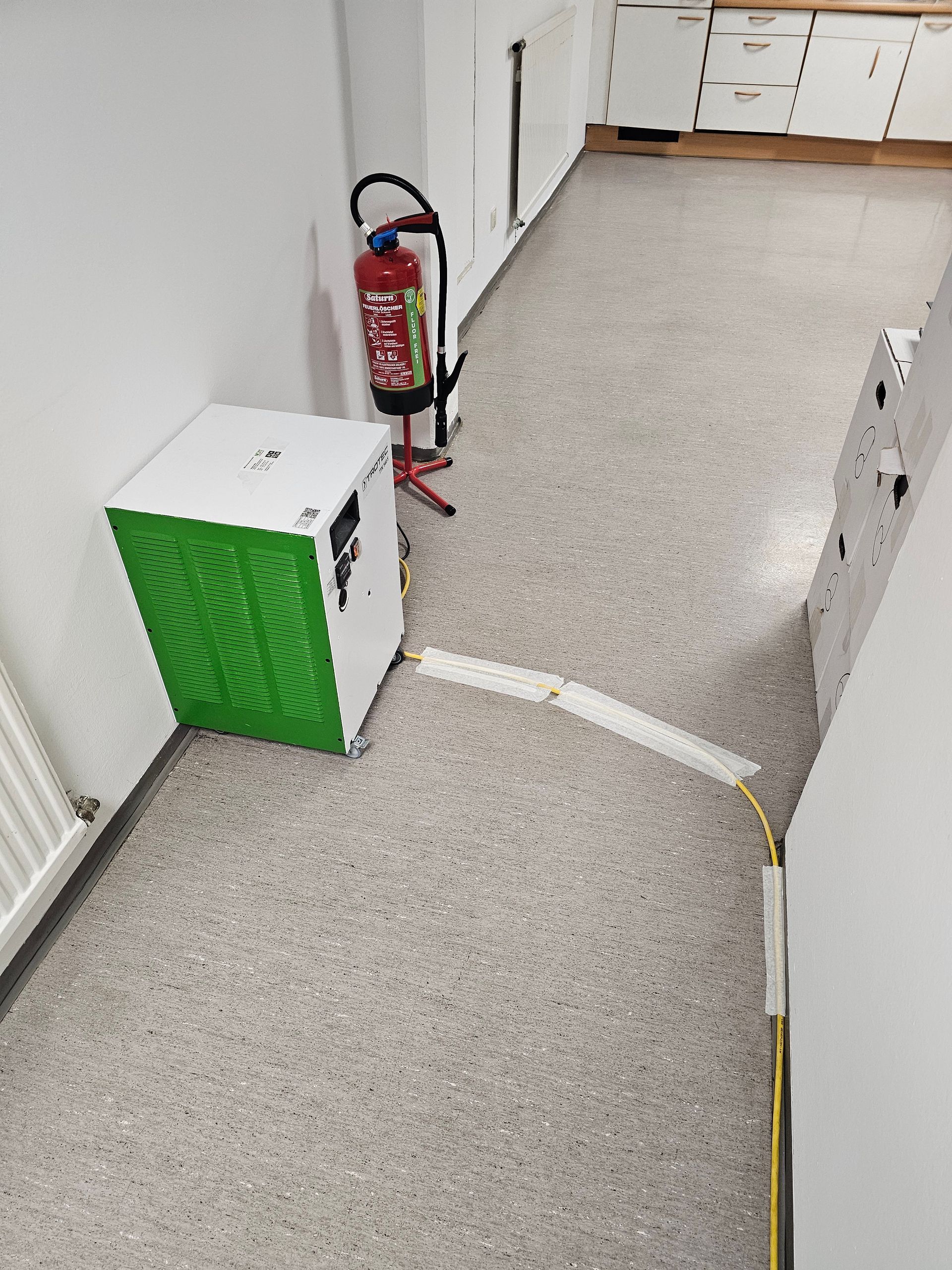 Neben einem Feuerlöscher steht ein weißes Gerät mit grüner Frontplatte; ein Kabel ist über eine Rampe auf dem Boden verdeckt.