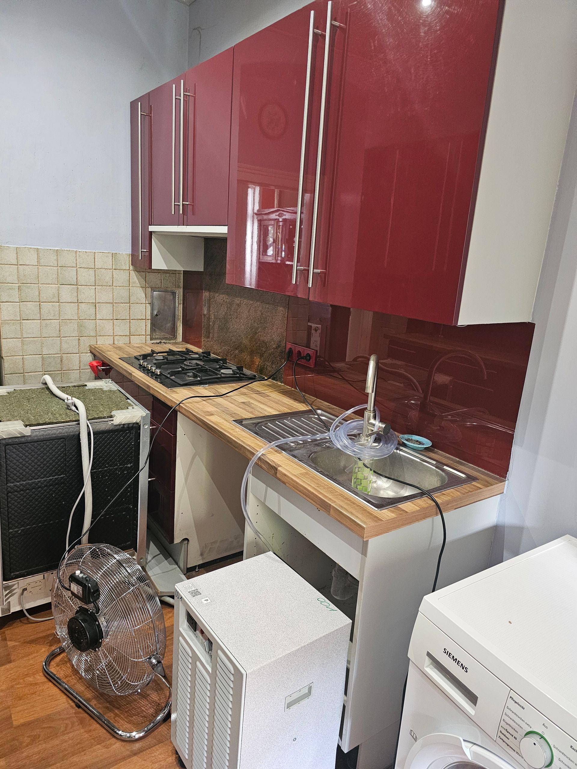 Eine Küche mit roten Oberschränken, einer Arbeitsplatte aus Holz, einem Spülbecken, einem Kochfeld und Sanitärinstallationen auf dem Boden.