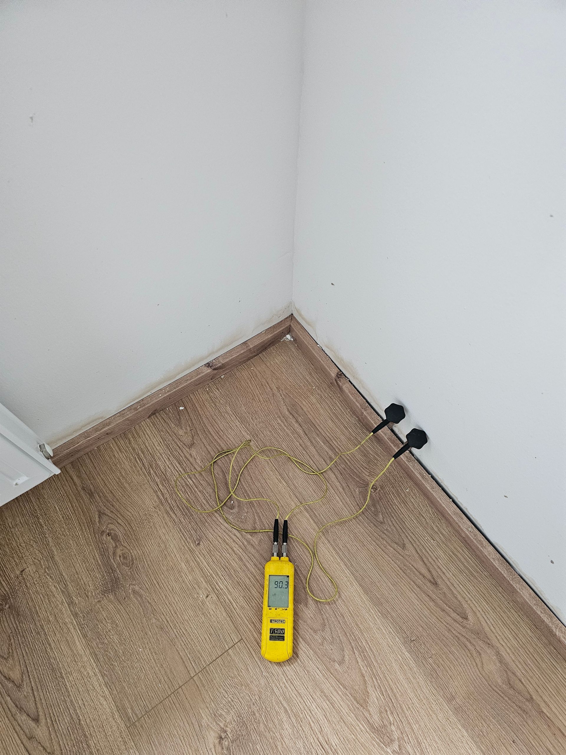 In einer Ecke des Zimmers, in der Nähe von Fußleisten mit dunklem Schimmelbefall, steht ein gelbes Feuchtigkeitsmessgerät auf einem Boden in Holzoptik.