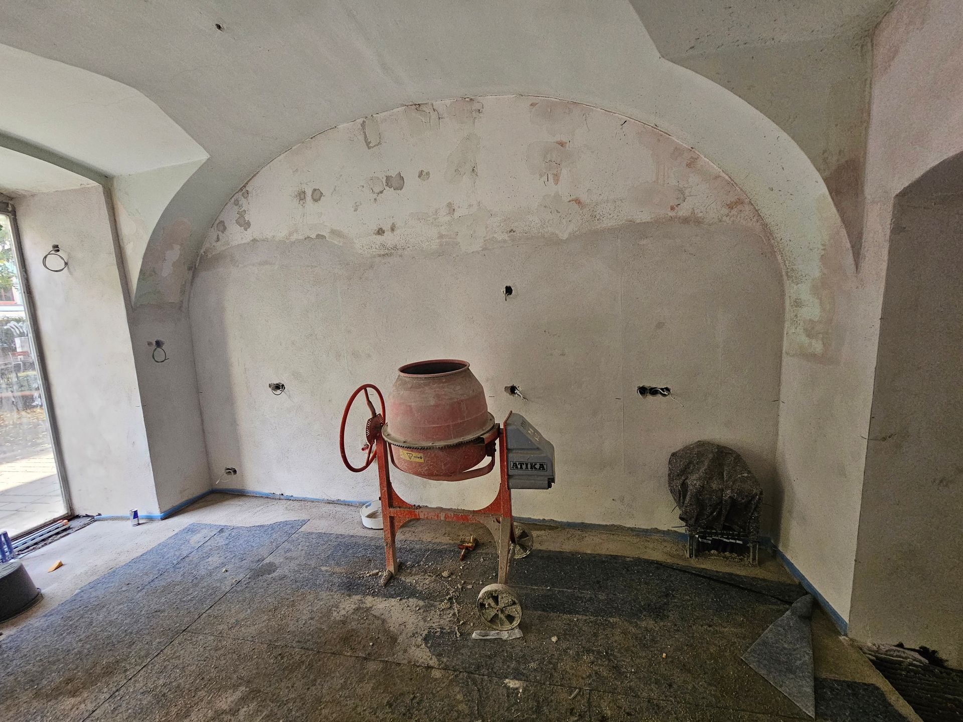 In der Mitte eines rustikalen Raumes mit gewölbter Decke und unfertigen Betonwänden und -boden steht ein Betonmischer.