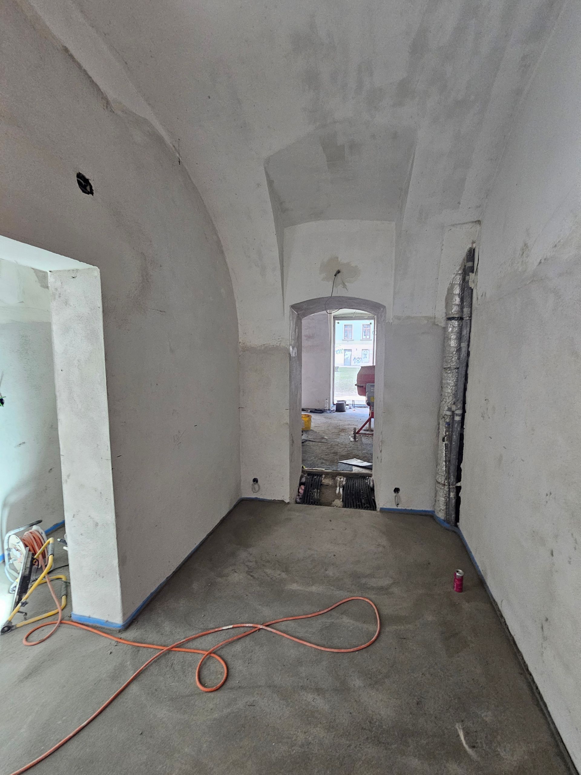 Ein Zimmer im Umbau mit unverputzten Wänden, einer gewölbten Decke und einer offenen Tür, die in einen anderen Raum führt.