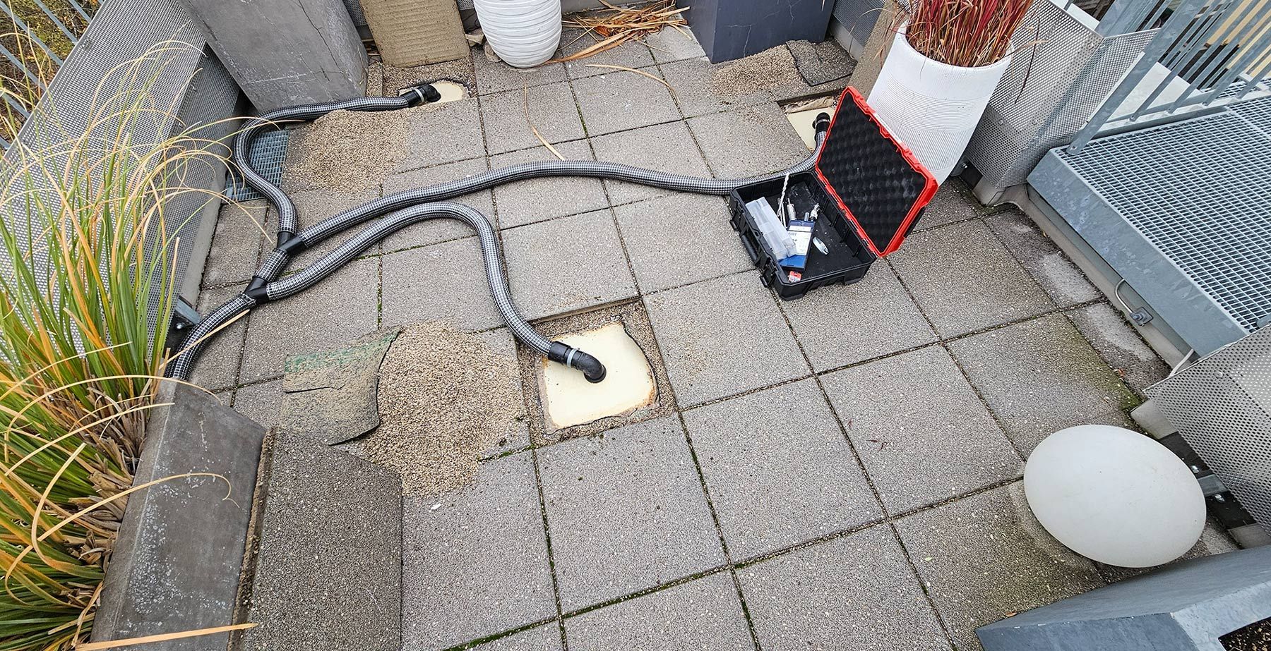 Ein schwarzer, offener Gerätekasten auf dem Terrassenboden ist über dicke, gerippte Schläuche mit einem quadratischen Abfluss im Pflaster verbunden.