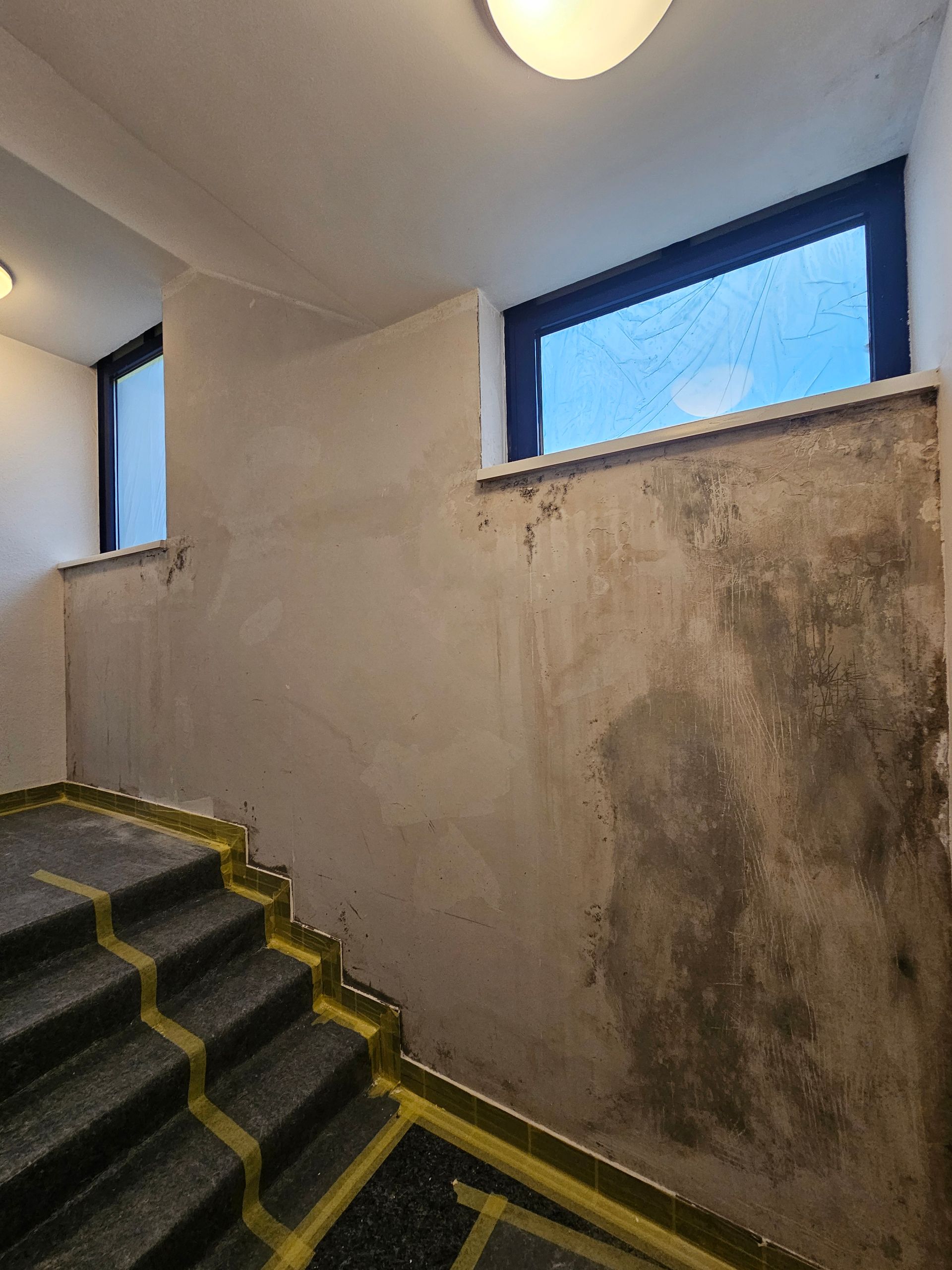 Treppenhaus mit unfertigen, ausgebesserten Putzwänden, gelbem Sicherheitsband auf den Stufen und zwei Fenstern.