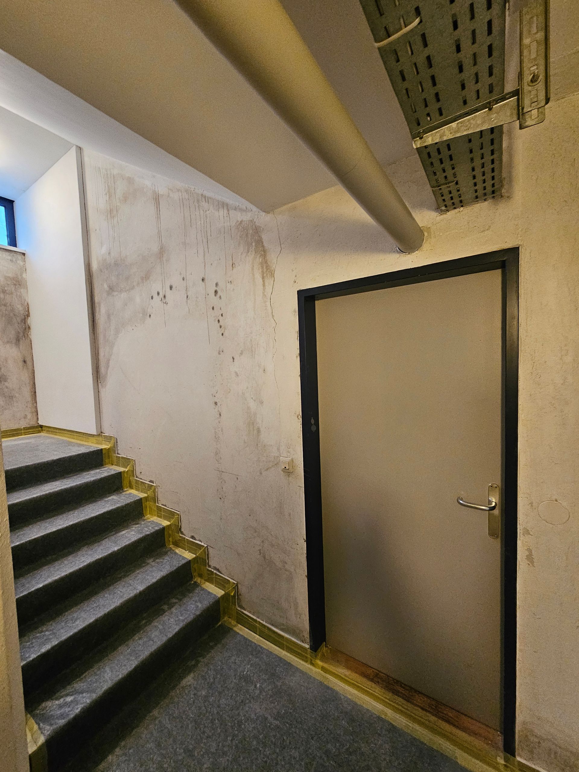 In einem Flur neben einer grauen Treppe steht eine graue Tür; die Wände sind fleckig und cremefarben, darüber verläuft eine Kabelrinne aus Metall.