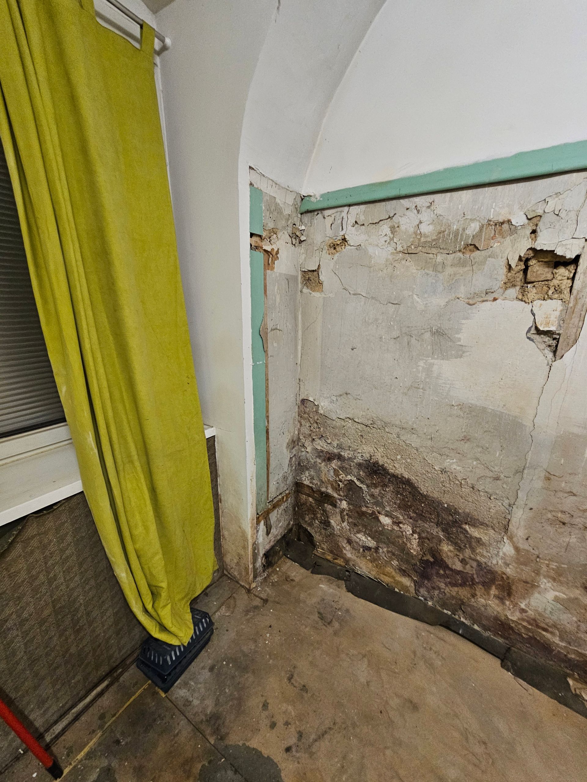 Eine Zimmerecke mit grünen Vorhängen neben einer beschädigten Wand, die Anzeichen von Feuchtigkeit, bröckelndem Putz und Mauerwerk aufweist.