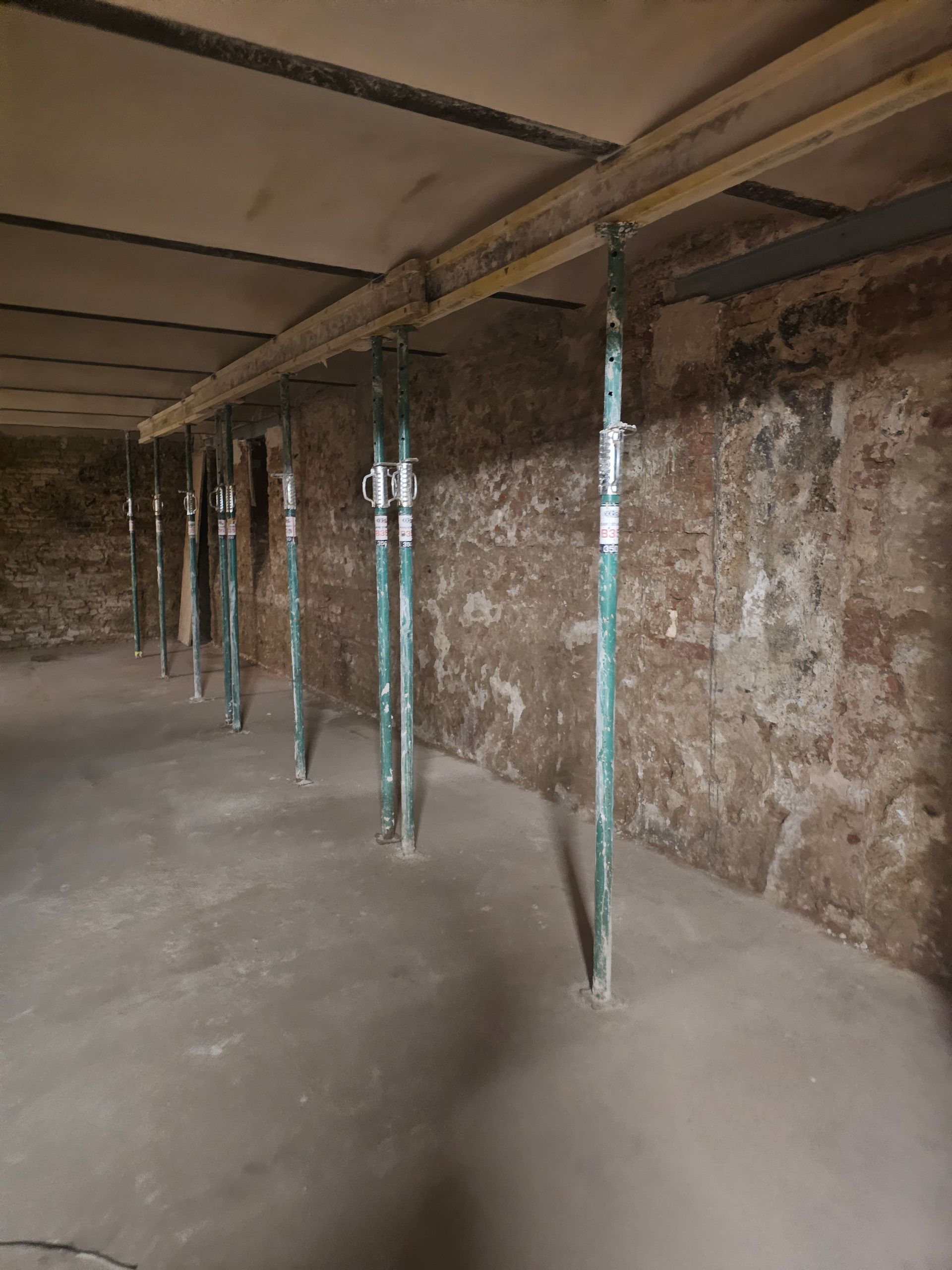 Im Inneren eines Kellers befinden sich mehrere vertikale Metallkonstruktionsstützen, die einen Holzbalken entlang einer groben Steinwand stützen.