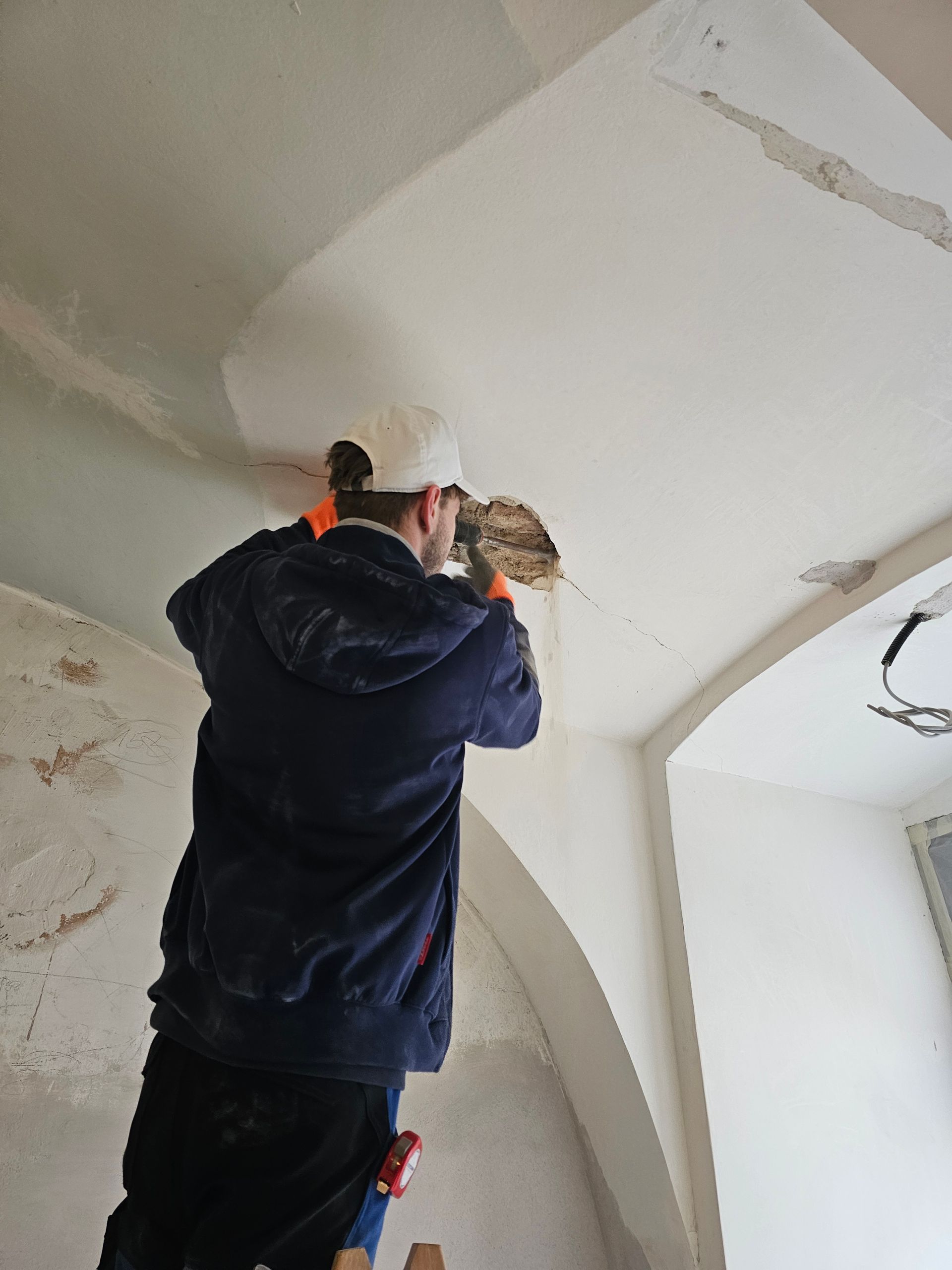 Ein Arbeiter auf einer Leiter verputzt eine gewölbte Decke in einem Raum, der renoviert wird.