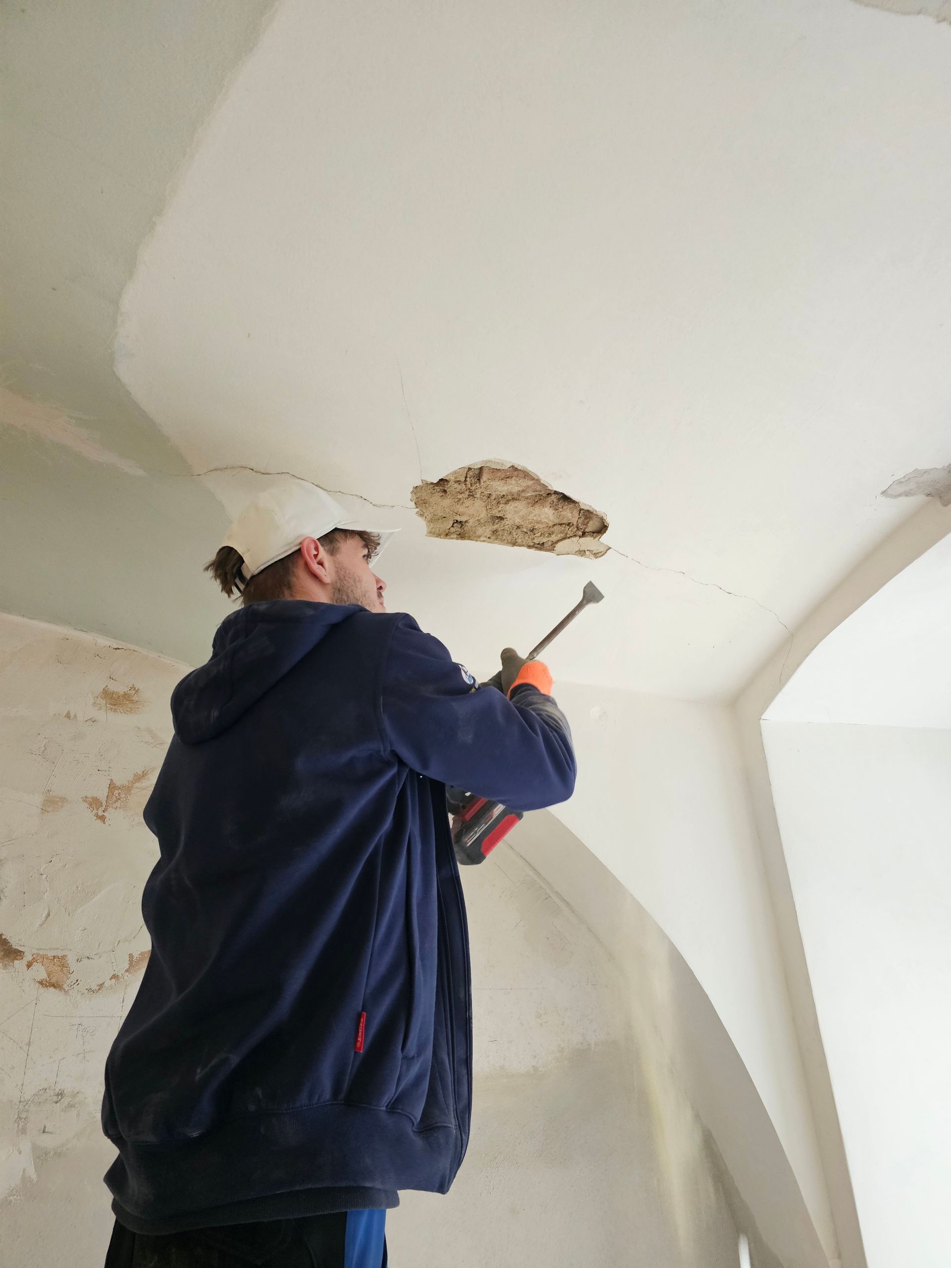 Eine Person in einem blauen Kapuzenpullover benutzt eine Kartuschenpistole, um eine beschädigte Stelle des weißen Putzes an einer gewölbten Decke auszubessern.