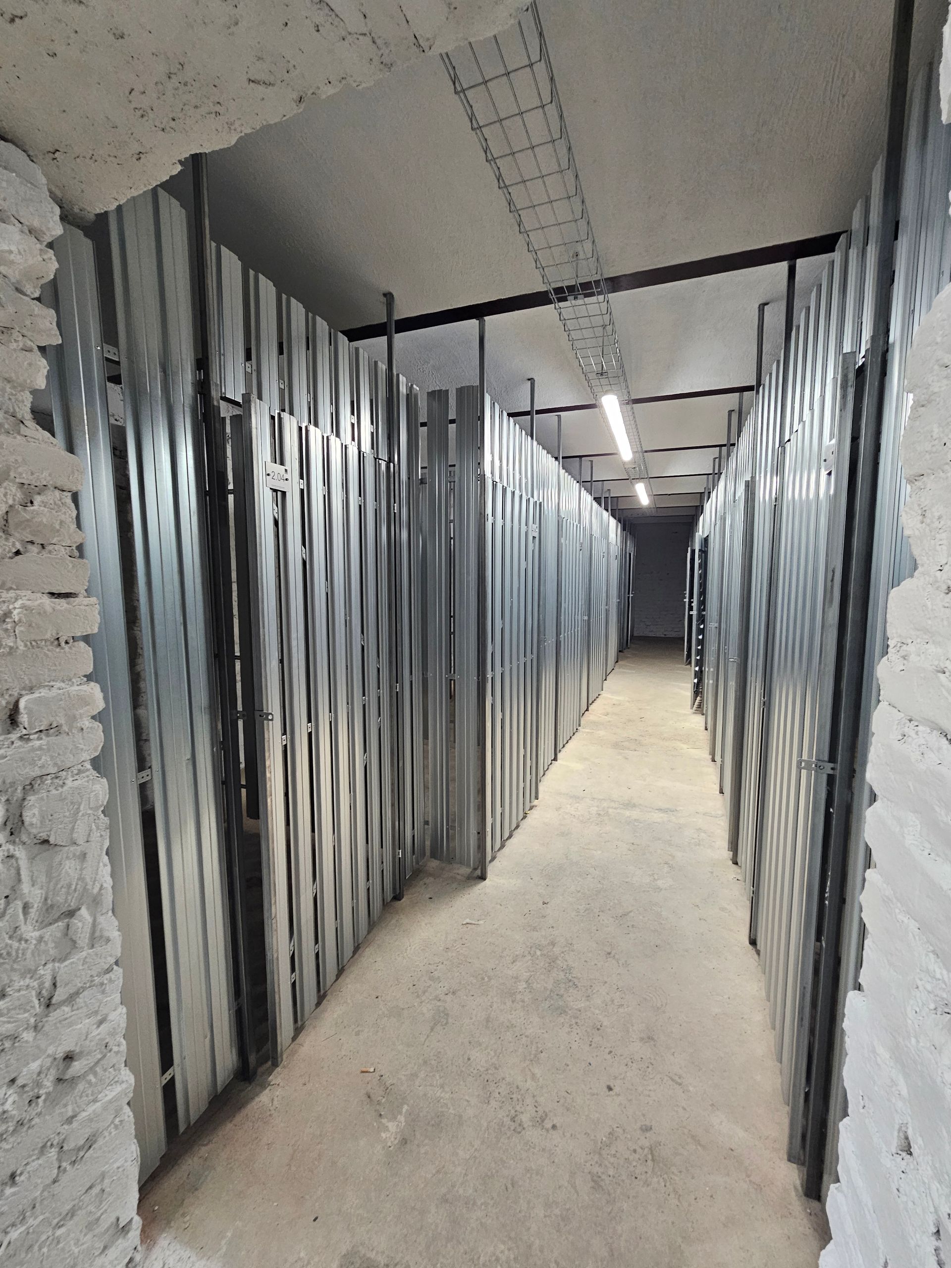 Ein schmaler Flur, gesäumt von hohen, vertikalen grauen Metallschränken und Türen, führt in einen dunkleren Raum.