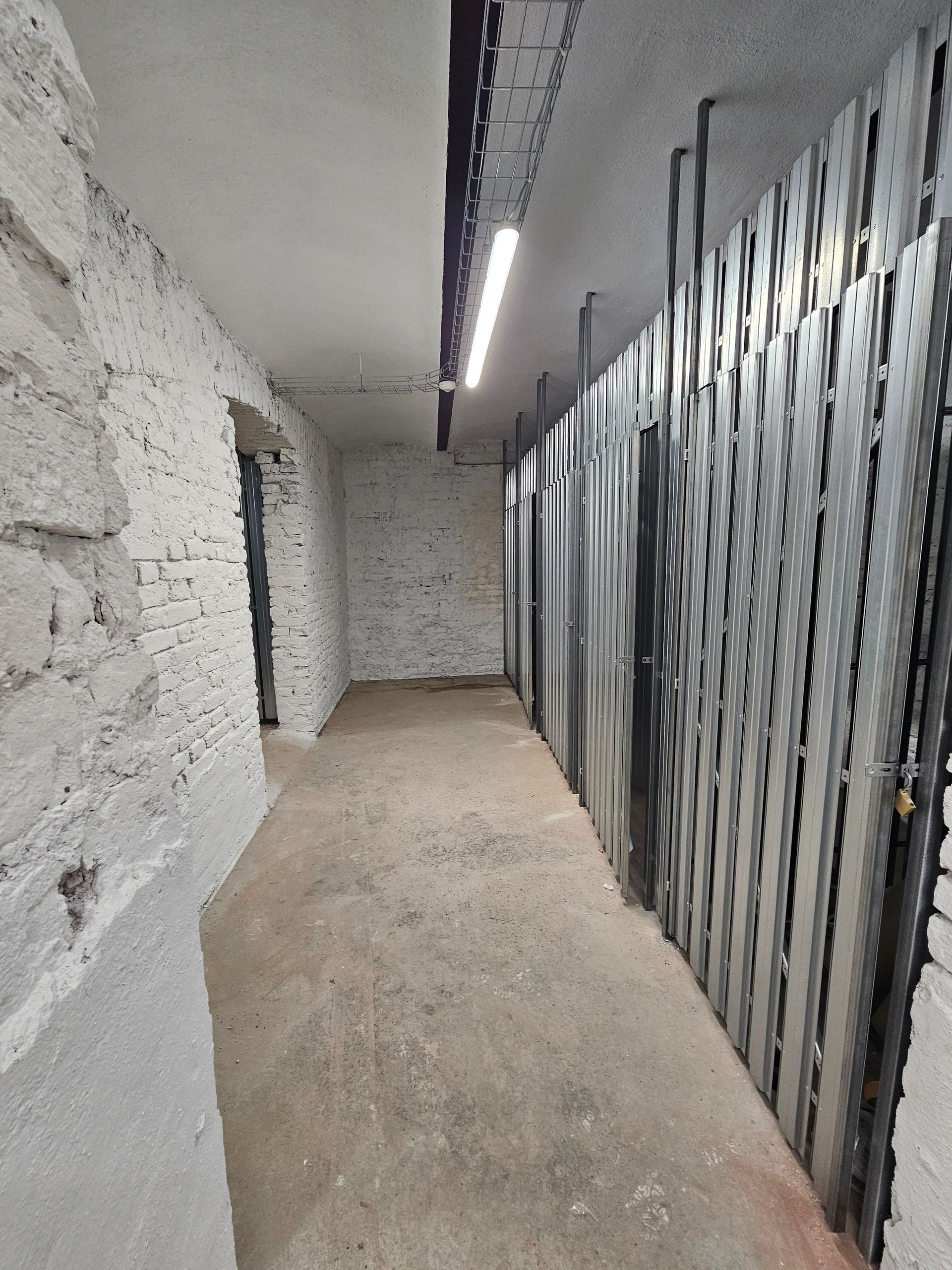 Ein schmaler, schwach beleuchteter Kellerflur mit einer weißen Backsteinwand auf der linken Seite und einer Reihe von Metallschränken auf der rechten Seite.