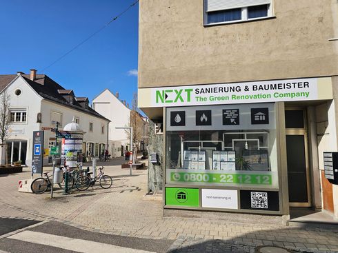 Ein Ladenlokal der Renovierungsfirma „NEXT“ in einer sonnigen Straße, vor dem Fahrräder geparkt sind.