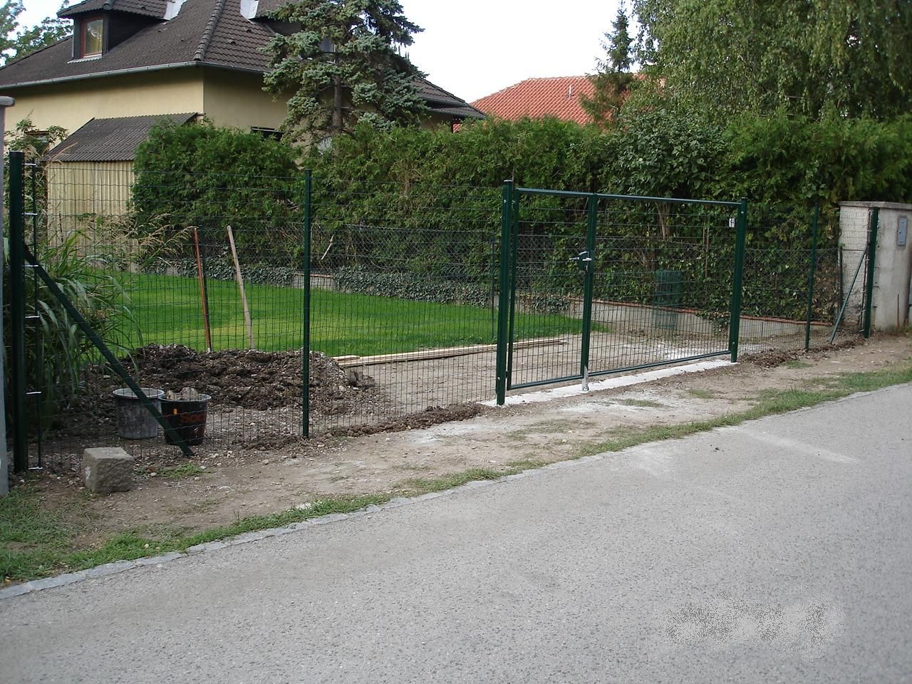 Ein dunkelgrüner Drahtgitterzaun mit Tor umgibt einen gepflegten Rasen und Garten neben einer gepflasterten Straße.