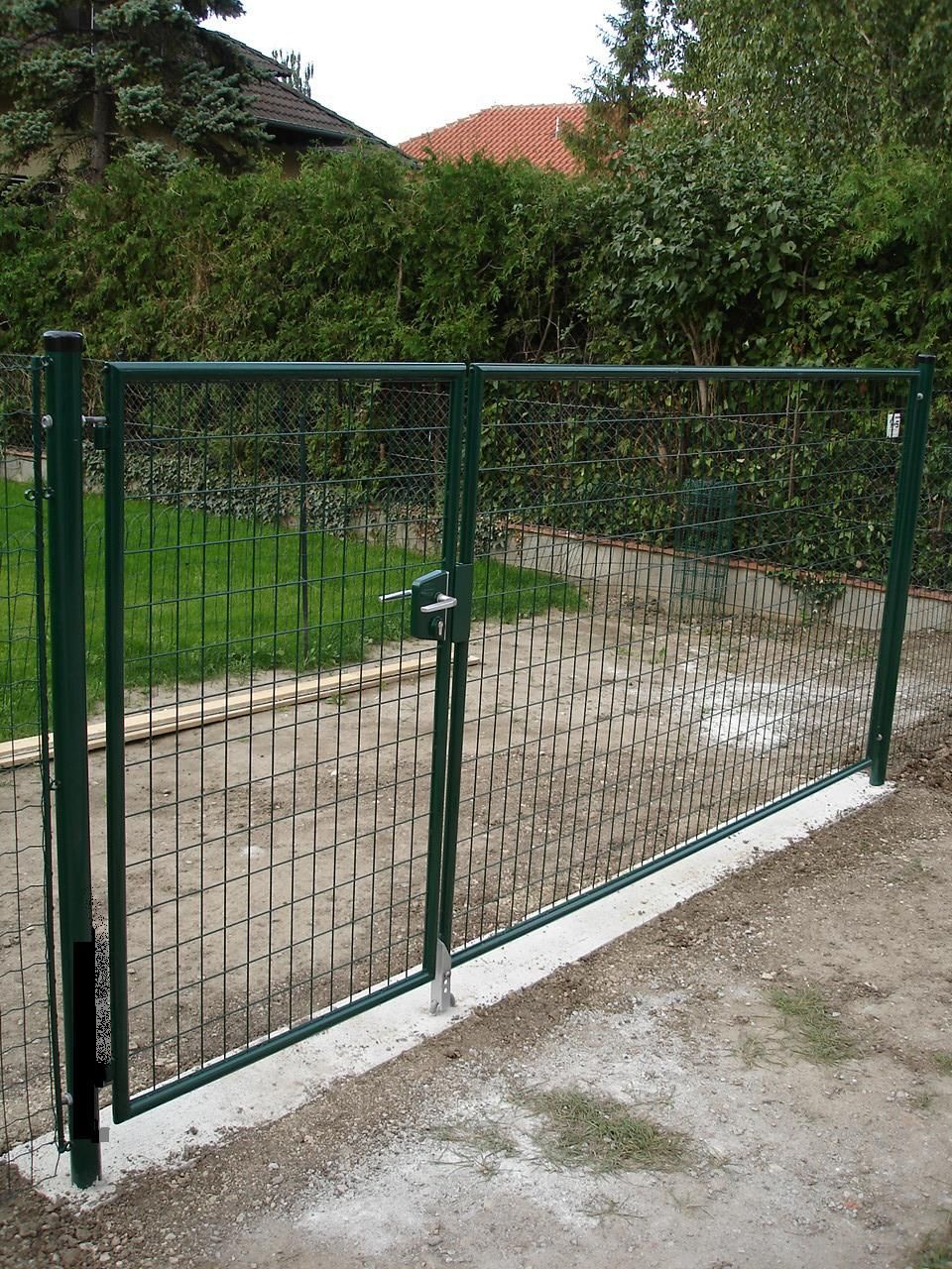 Ein dunkelgrünes Metall-Fußgängertor, eingefasst in einen Drahtgitterzaun, installiert auf einem Betonbordstein vor einer Hecke.