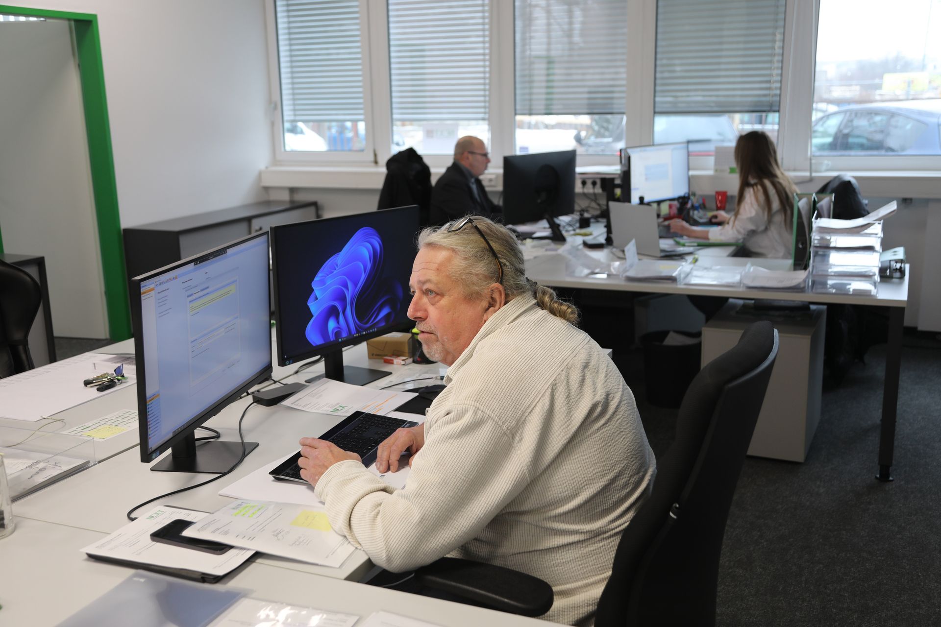 Ein Mann arbeitet an einem Computer in einem Büro, im Hintergrund arbeiten zwei Kollegen an Schreibtischen in der Nähe der Fenster.