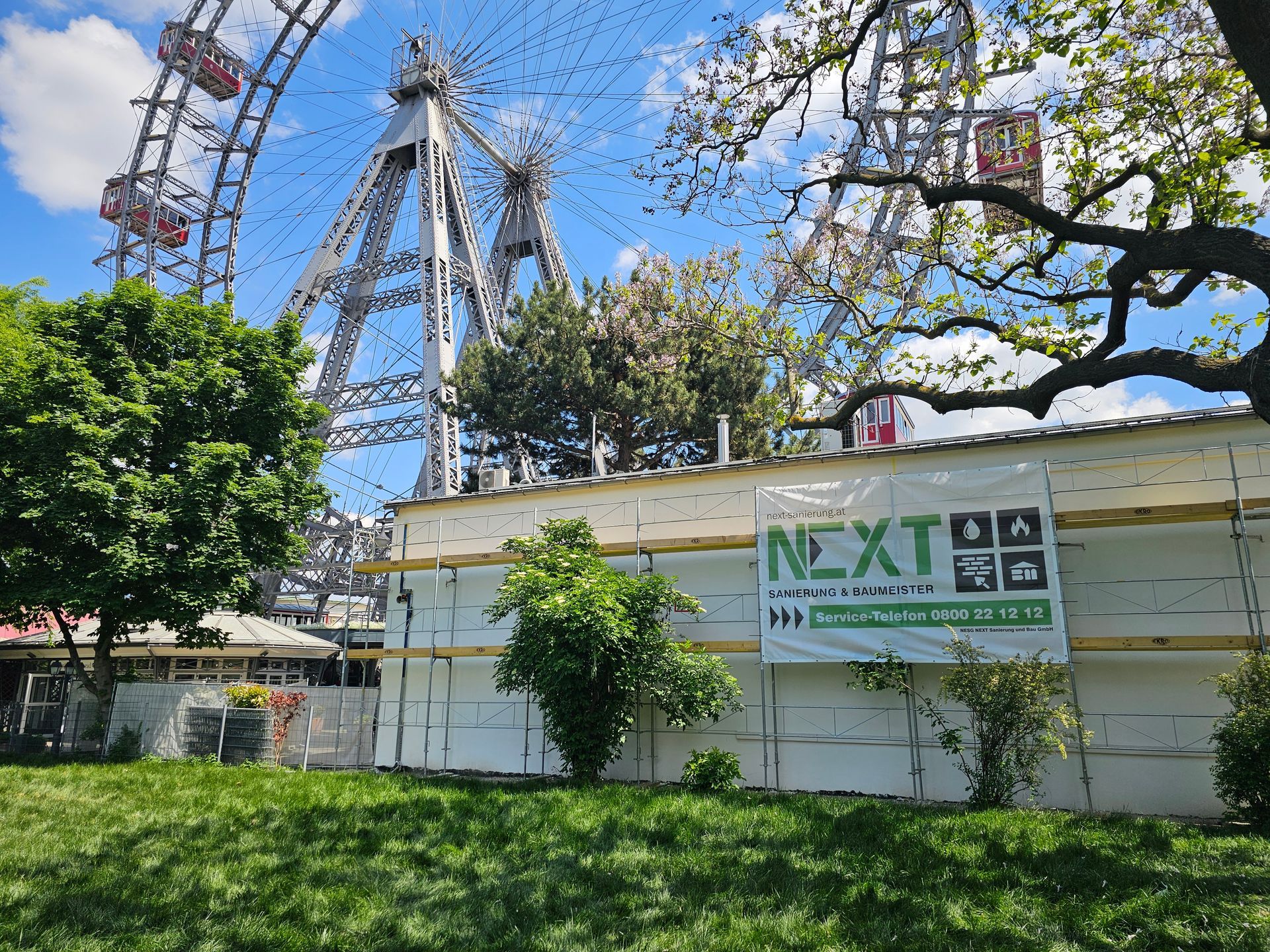 Das Wiener Riesenrad in Wien steht hinter einer weißen Bauwand mit einem grünen Baustellenschild „Next“.