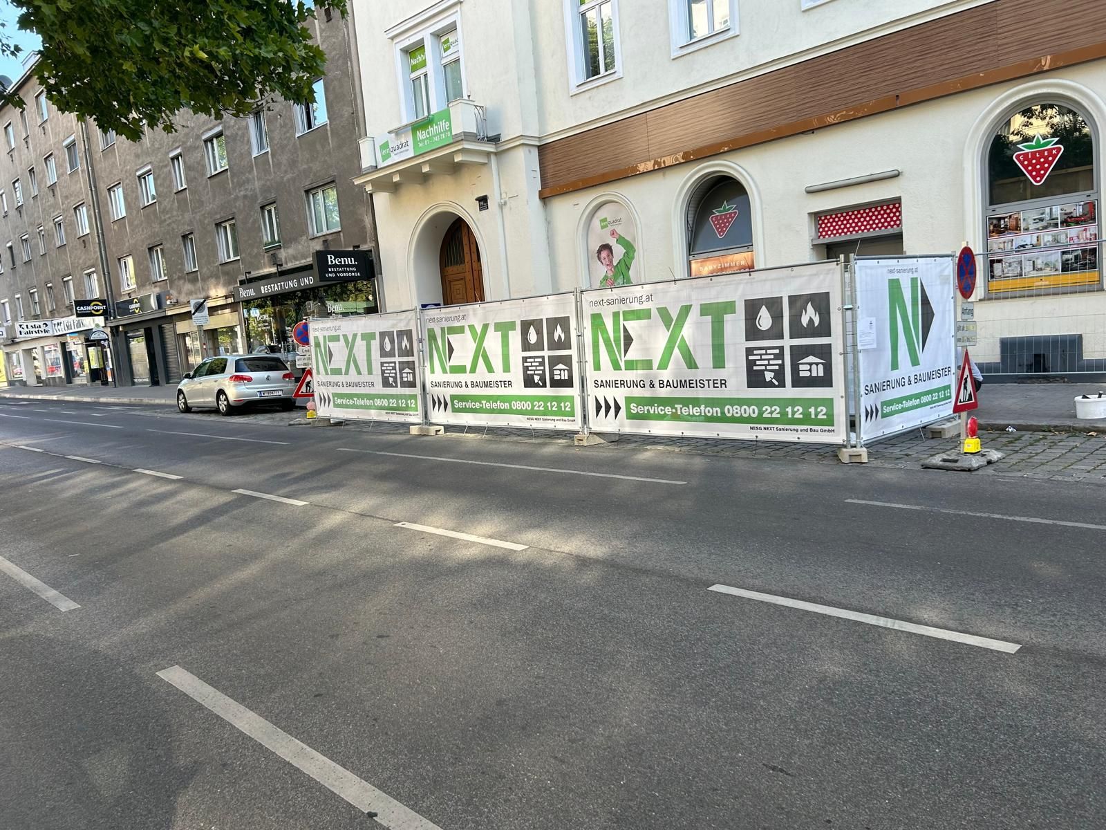 Bauzaun mit der Aufschrift „NEXT“ vor einem mehrstöckigen Gebäude an einer Straße in der Stadt.