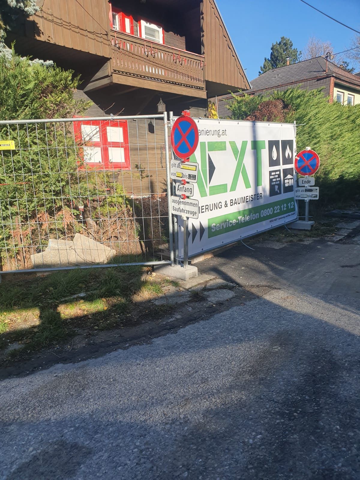 Ein Bauzaun mit einem großen „NEXT“-Banner und zwei Parkverbotsschildern vor einem Haus an einem sonnigen Tag.