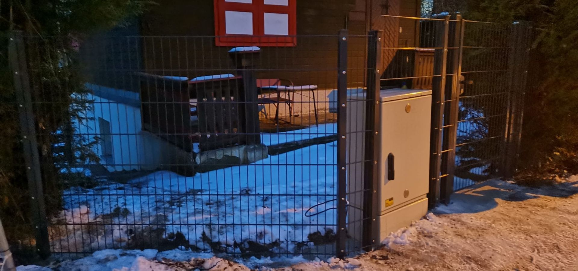 Ein grauer Metallschrank steht außerhalb eines Drahtzauns vor einem dunklen Gebäude mit rotgerahmten Fenstern im Schnee.