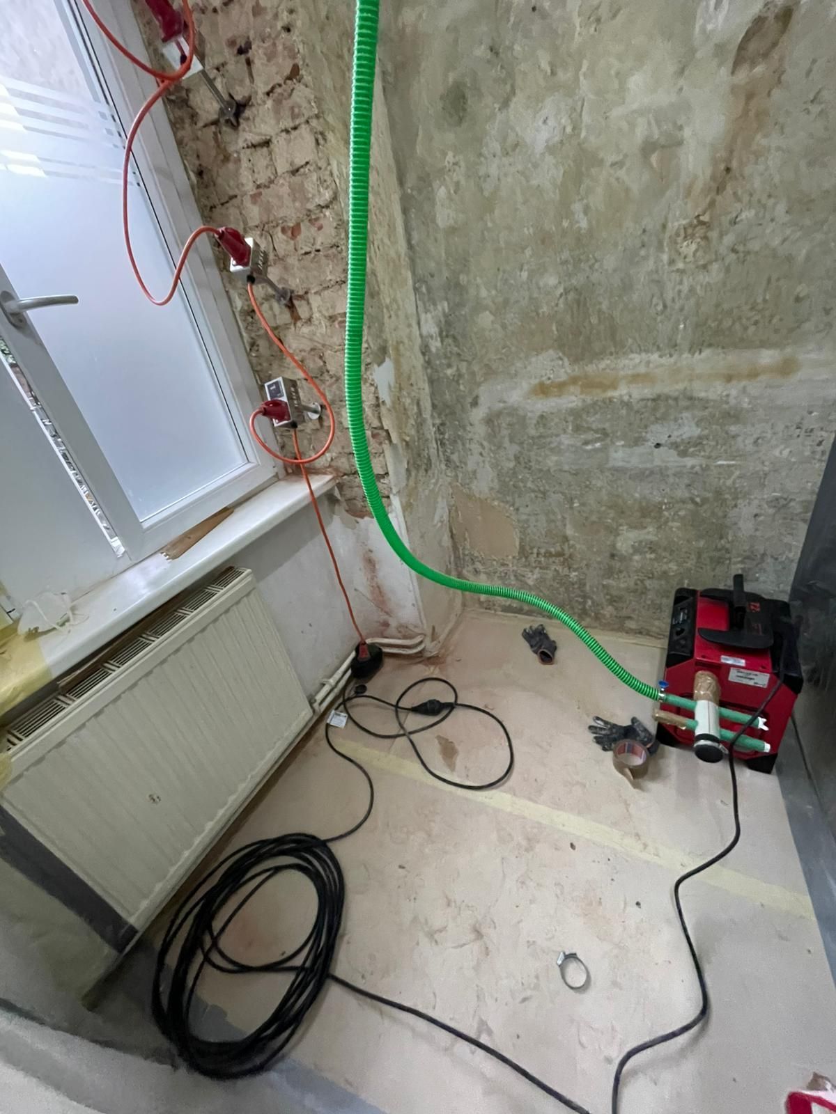 Auf einer Baustelle ist eine rote Maschine zu sehen, die mit einem grünen Schlauch verbunden ist. Schwarze Kabel liegen auf dem Boden neben einem Heizkörper.