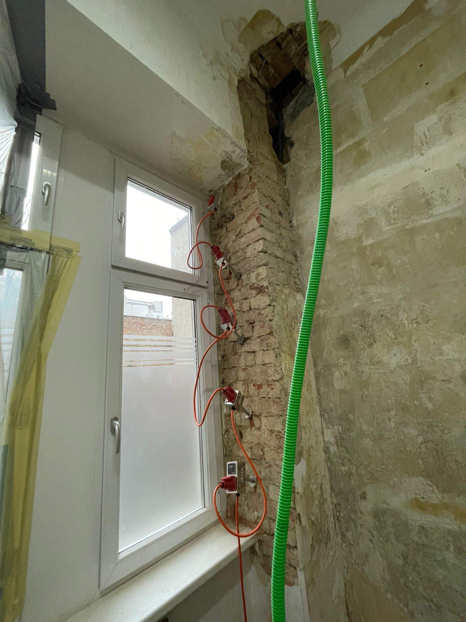 Eine im Bau befindliche Innenwand mit freiliegendem Ziegelstein, einer Steckdose, herabhängenden Kabeln und einem nahegelegenen Fenster.