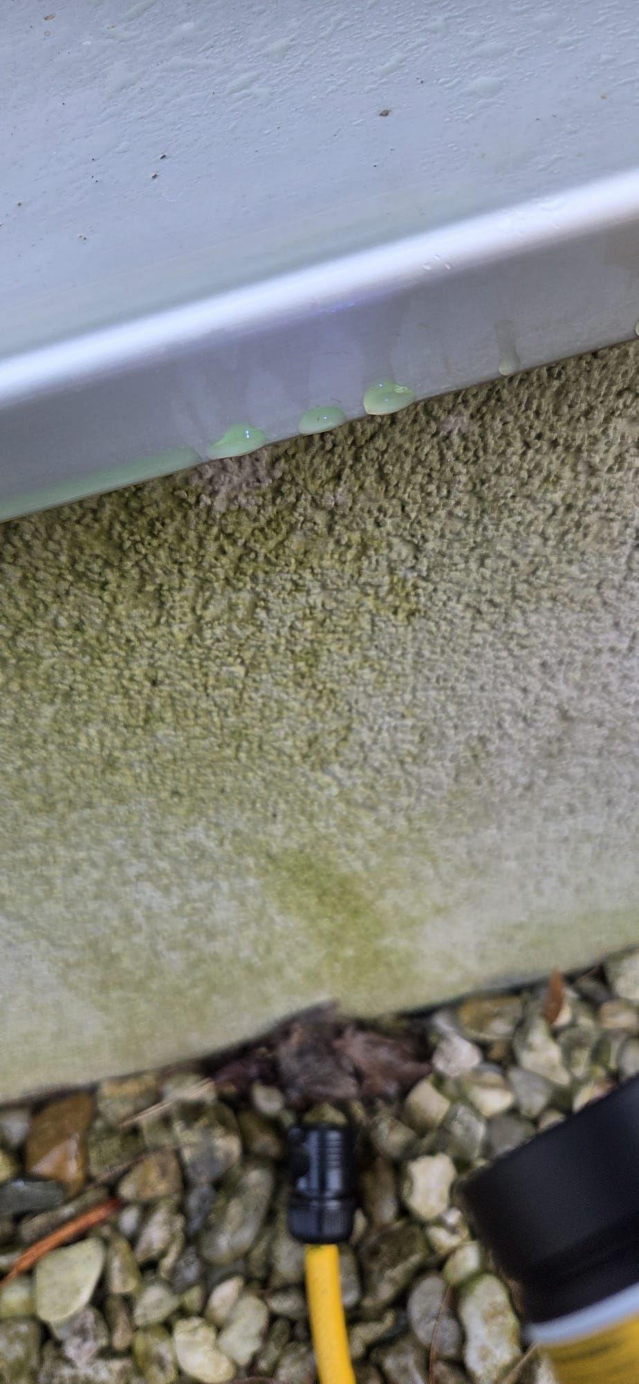 Wasser tropft von einer metallischen Dachkante auf eine strukturierte, algenbewachsene Wand über einer Bodenschicht aus Kieselsteinen und einer gelben Schnur.