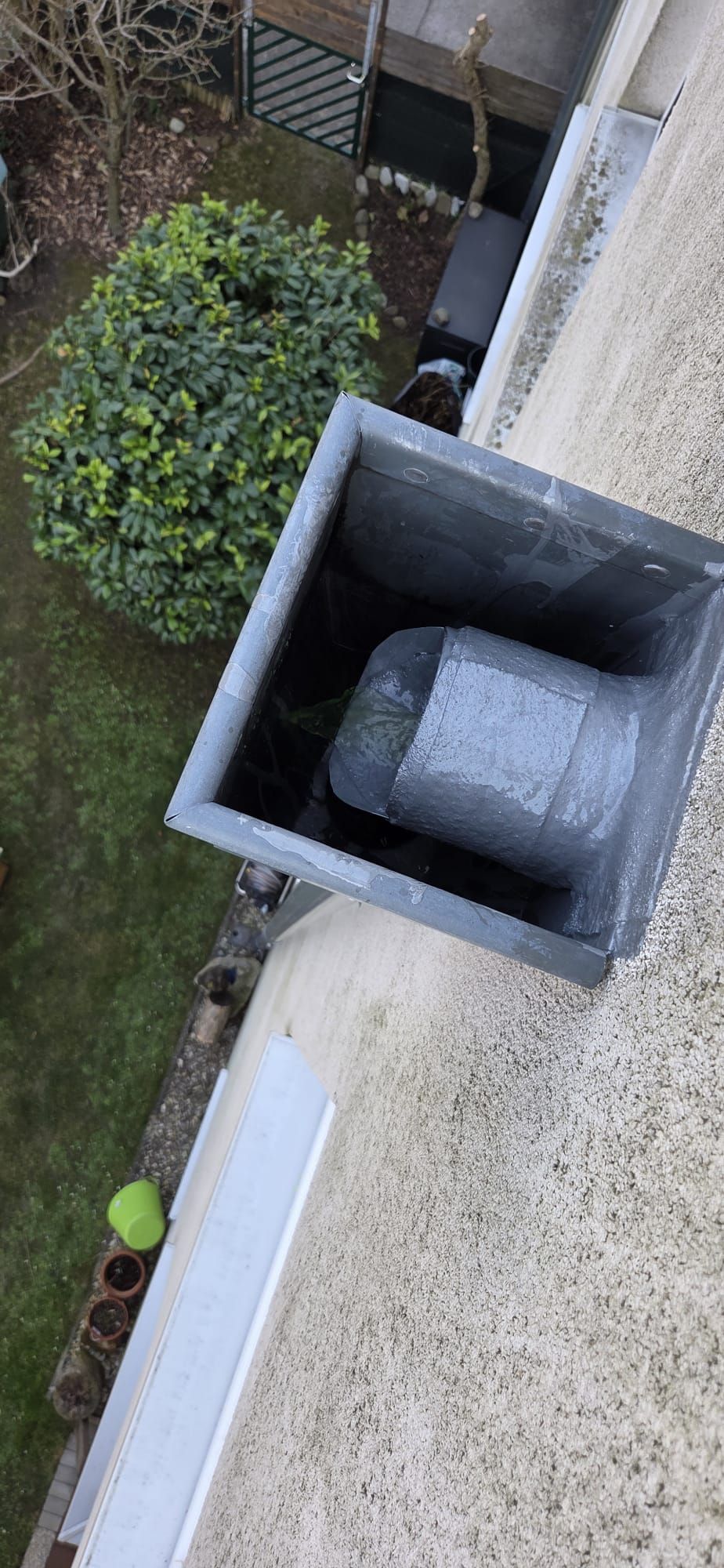 Draufsicht auf eine quadratische, graue Schornsteinöffnung auf einem Dach mit einem runden, grauen Lüftungsrohr im Inneren.