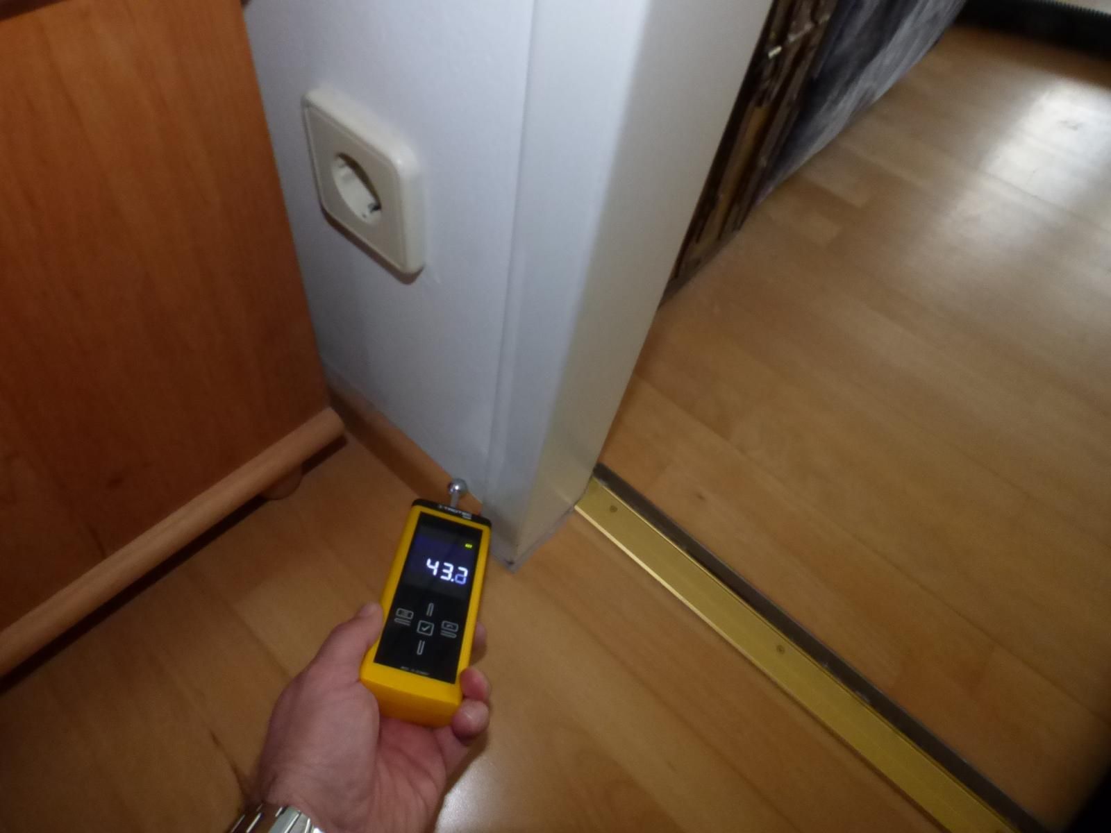 Eine Hand hält ein gelbes digitales Feuchtigkeitsmessgerät an einen weißen Türrahmen in der Nähe einer Steckdose und eines Hartholzbodens.