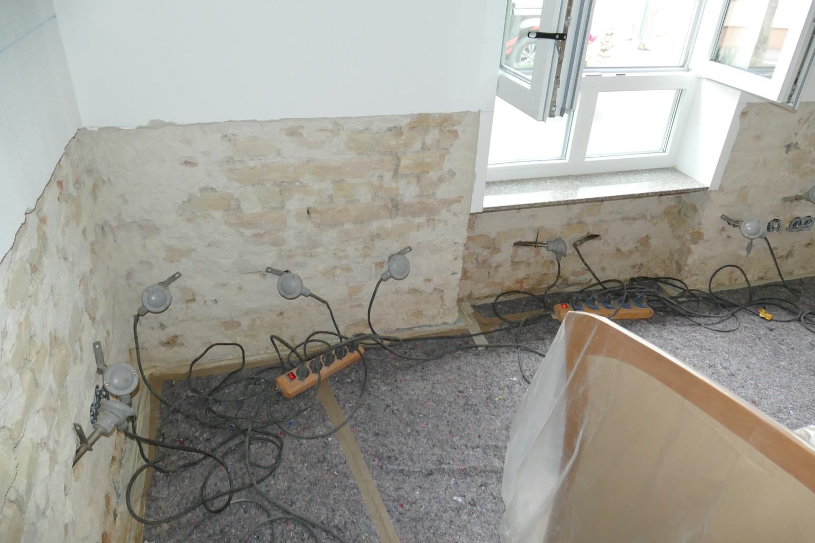 Ein Raum, in dem gerade eine Wandreparatur durchgeführt wird, mit mehreren Arbeitsleuchten, die an Steckdosenleisten auf dem Boden in der Nähe eines offenen Fensters angeschlossen sind.