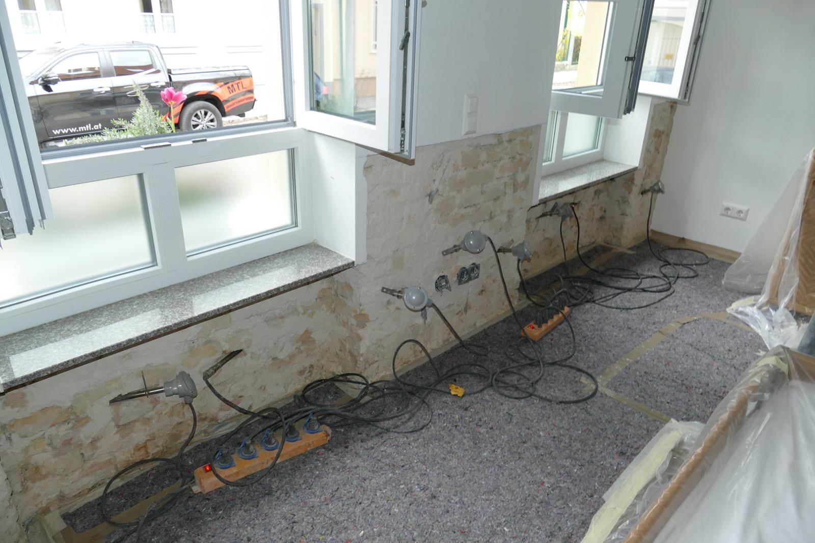 Ein Innenraum im Umbauprozess mit freiliegenden Betonwänden, Stromkabeln und Steckdosenleisten auf dem Boden.