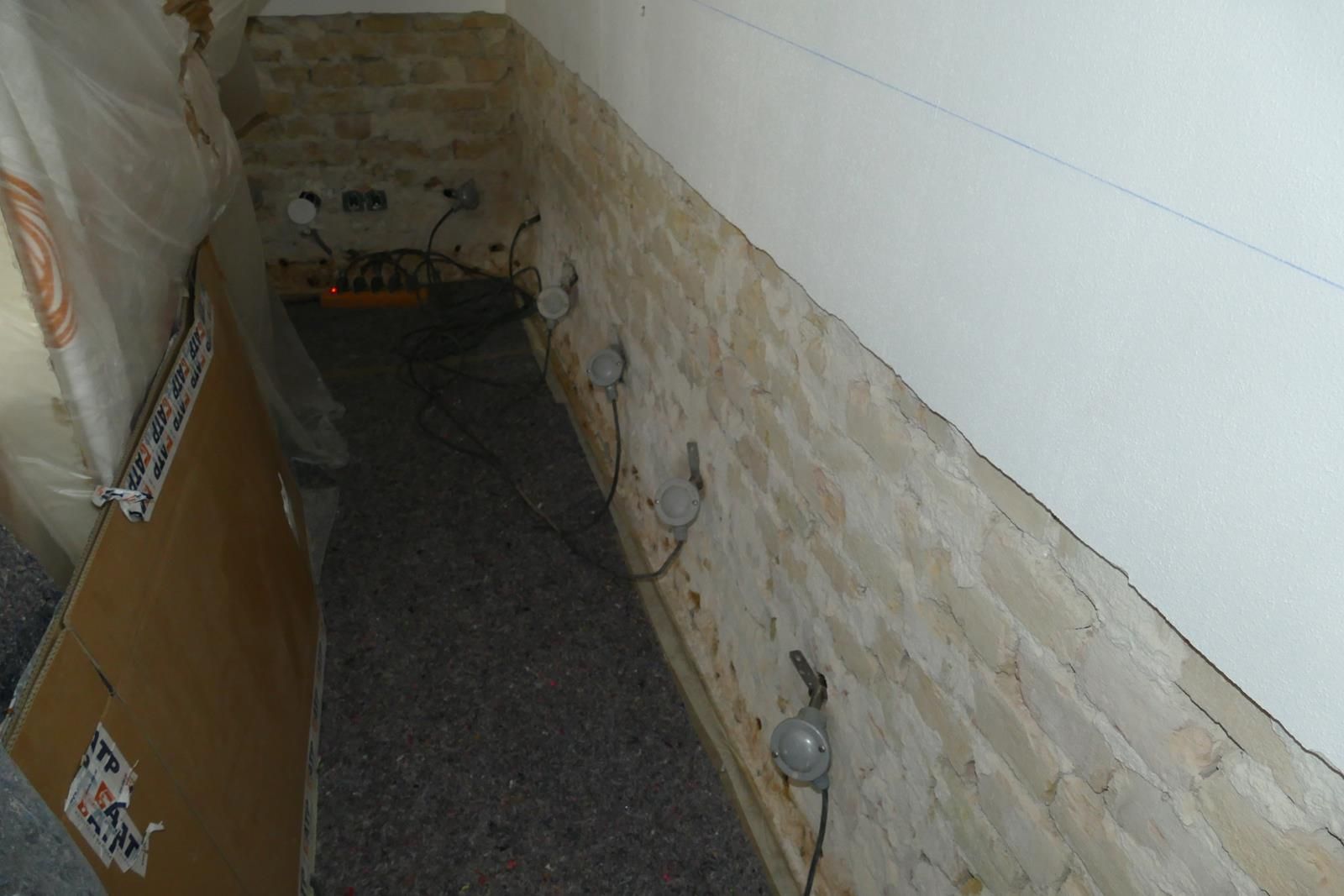 Glühbirnen sind an einer Wand in einem schmalen, schwach beleuchteten Raum mit Kiesboden und freiliegendem Ziegelstein angebracht.