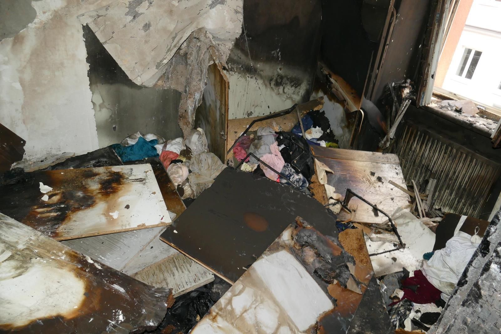 Ein Zimmer nach einem Brand, mit geschwärzten, mit Trümmern bedeckten Möbeln, versengten Wänden und Überresten von Haushaltsgegenständen.