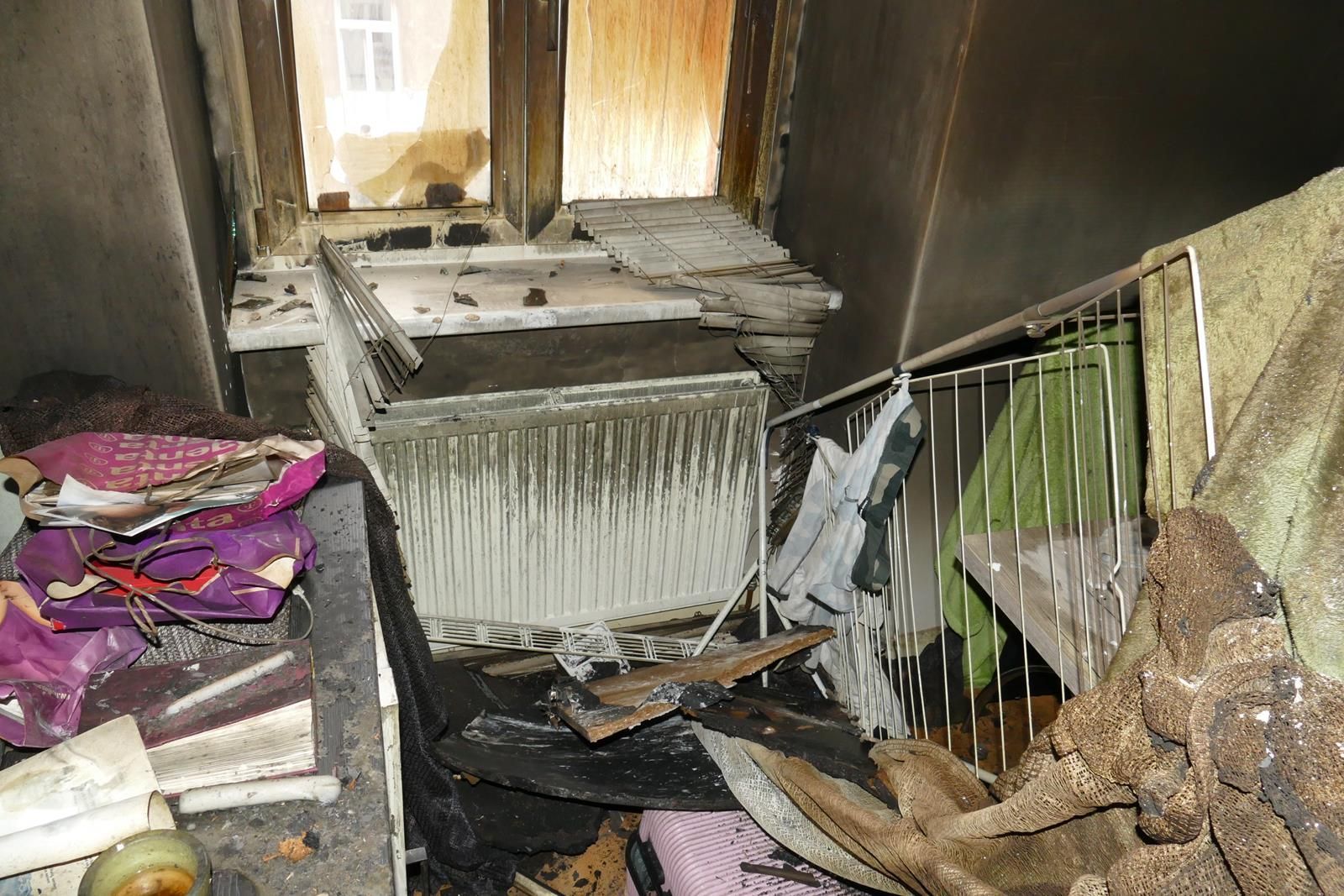 Ein vollgestelltes, durch einen Brand beschädigtes Zimmer mit einem Heizkörper unter einem Fenster, bedeckt mit Ruß, Schutt und verbrannten Gegenständen.