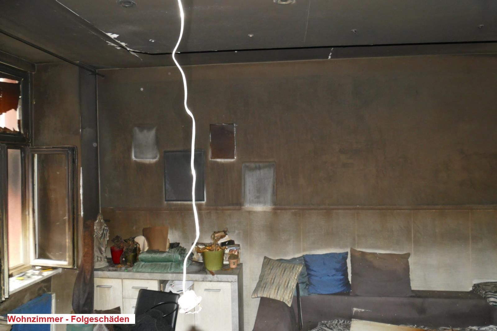 Ein durch Feuer beschädigtes Zimmer mit rußbefleckten Wänden und Decke, einem Sofa mit Kissen und einer freiliegenden Lampe.