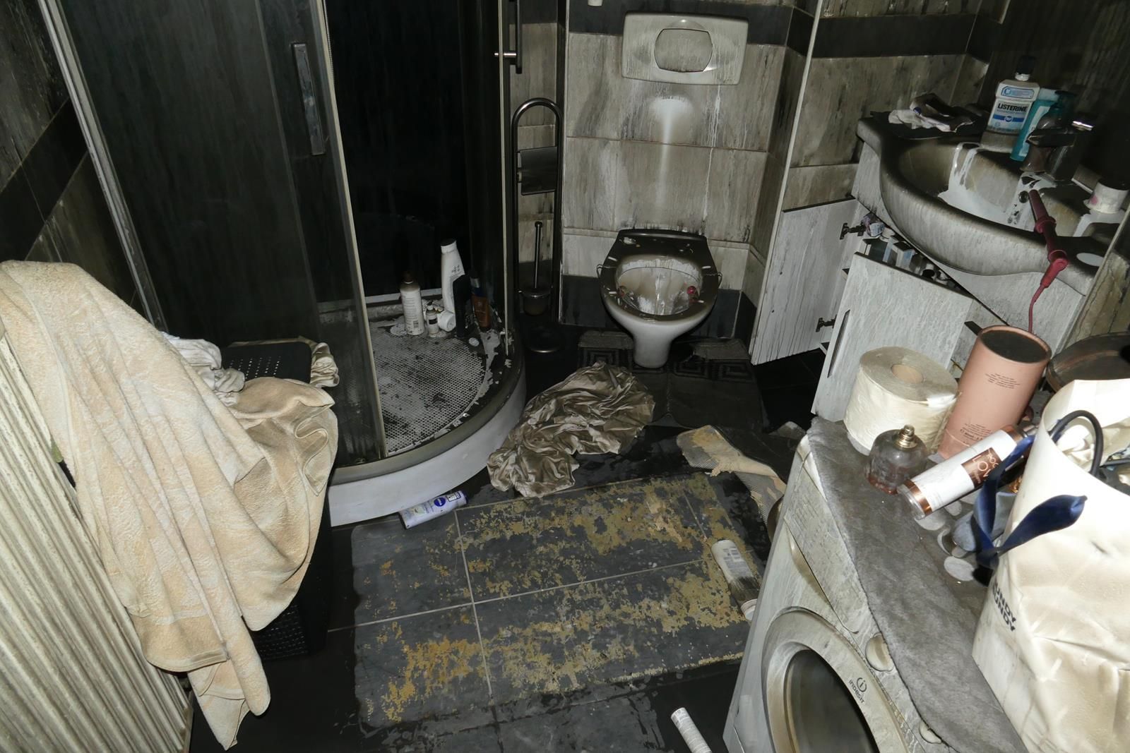 Ein unordentliches, schlecht beleuchtetes Badezimmer mit Toilette, Dusche, Waschbecken und verstreuter Wäsche auf dem dunklen Boden.