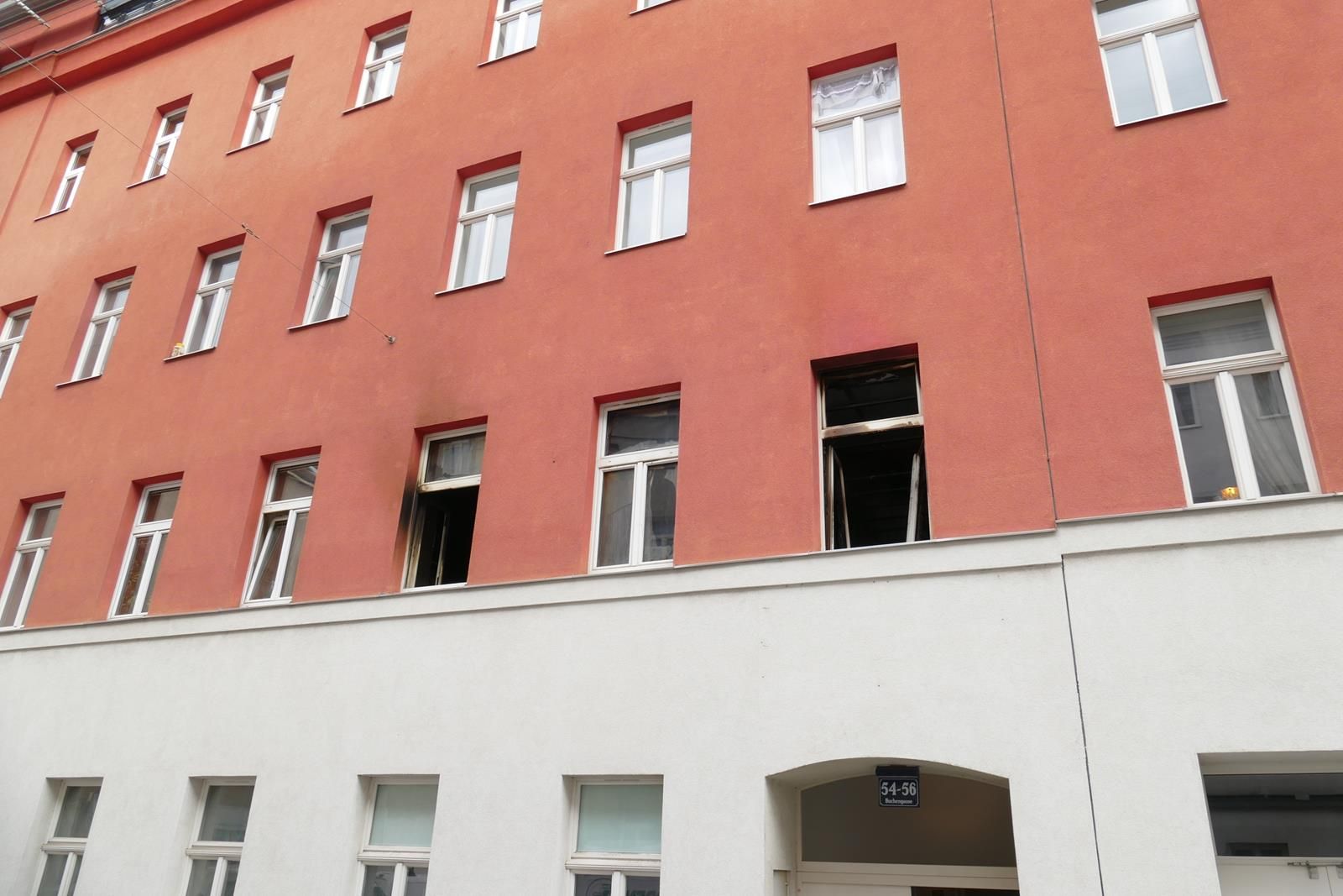Ein mehrstöckiges Gebäude mit einer terrakottafarbenen oberen Fassade und weißem Erdgeschoss, an dessen zwei Fenstern Brandschäden sichtbar sind.