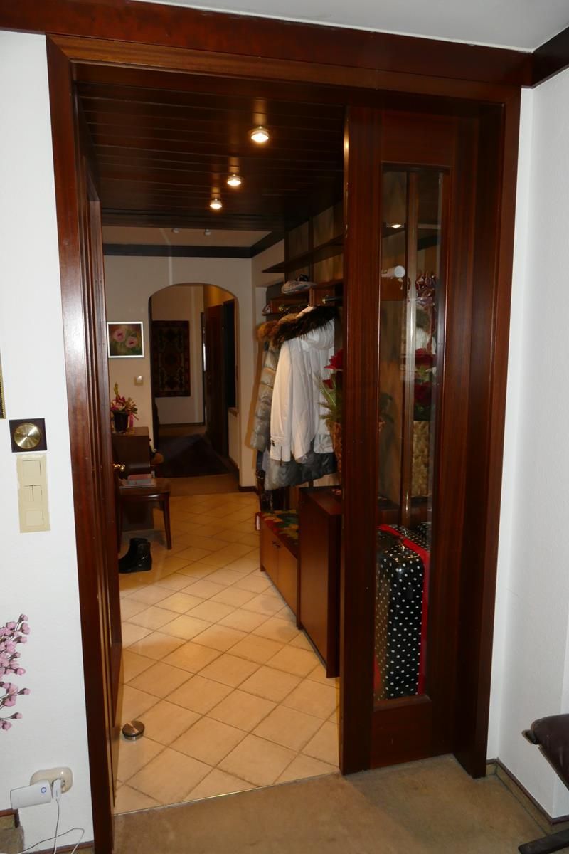 Eine dunkle, holzgerahmte Schiebetür mit Glasscheibe führt in einen Flur mit gefliestem Boden und hängender Kleidung.