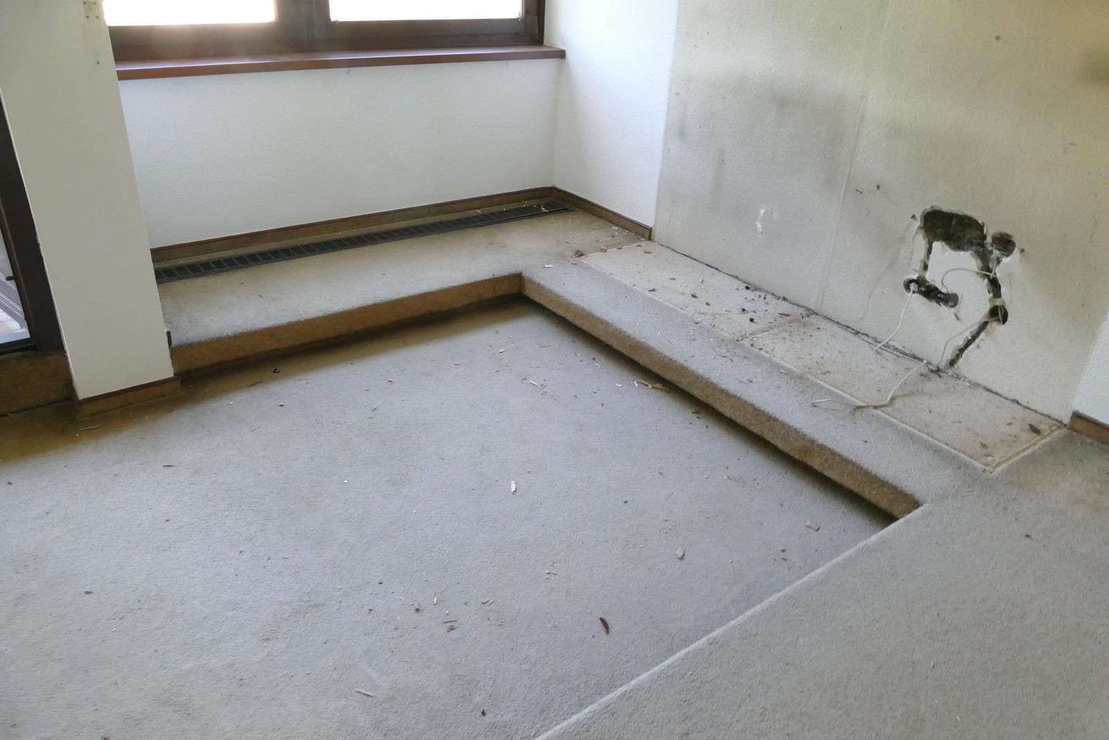 Ein L-förmiger, abgesenkter Bodenbereich in einem Raum mit betonähnlicher Oberfläche, einem Fenster und freiliegenden Wandleitungen.