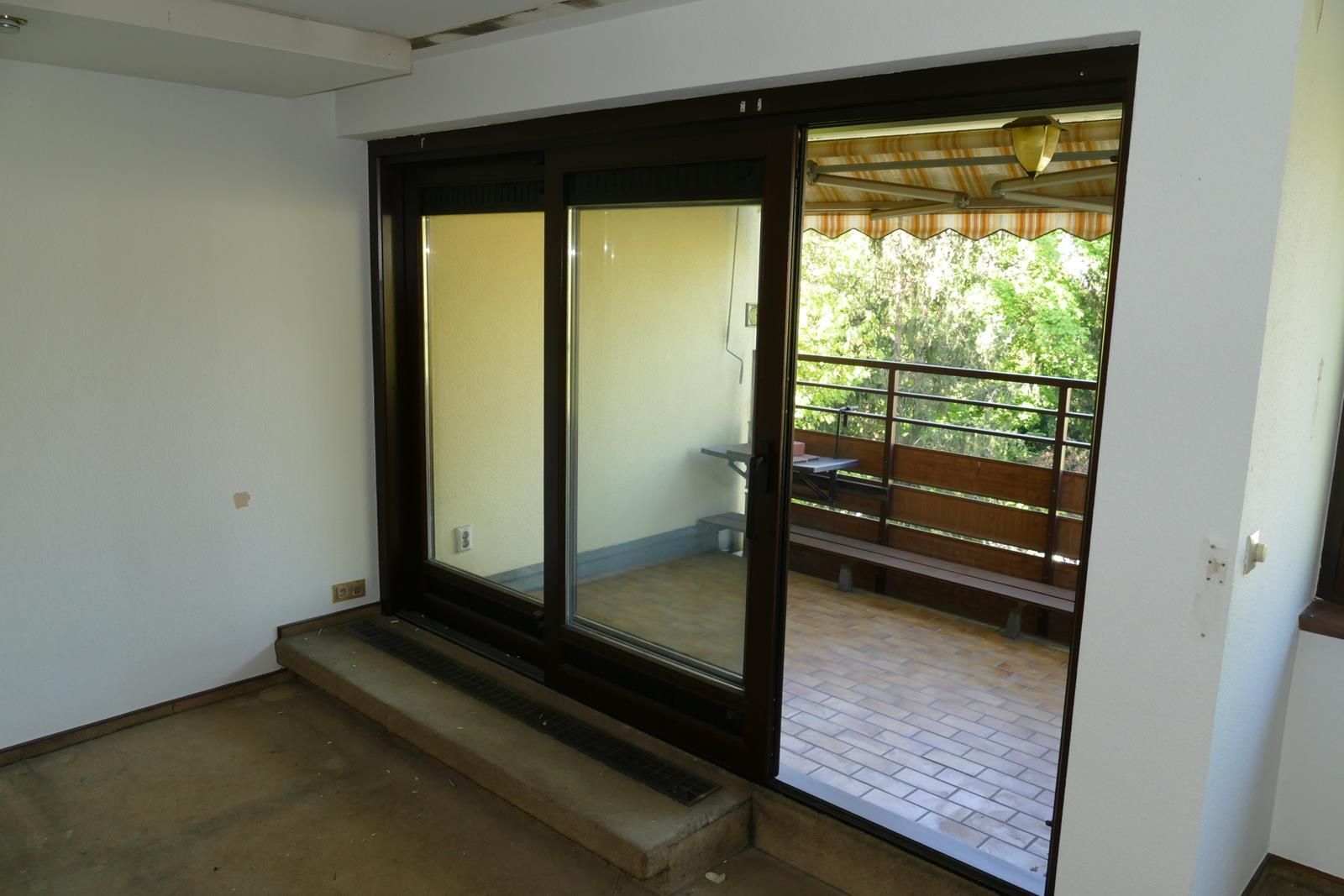 Eine dunkel gerahmte Glasschiebetür öffnet sich zu einem Balkon mit braunem Holzgeländer und Ziegelboden.