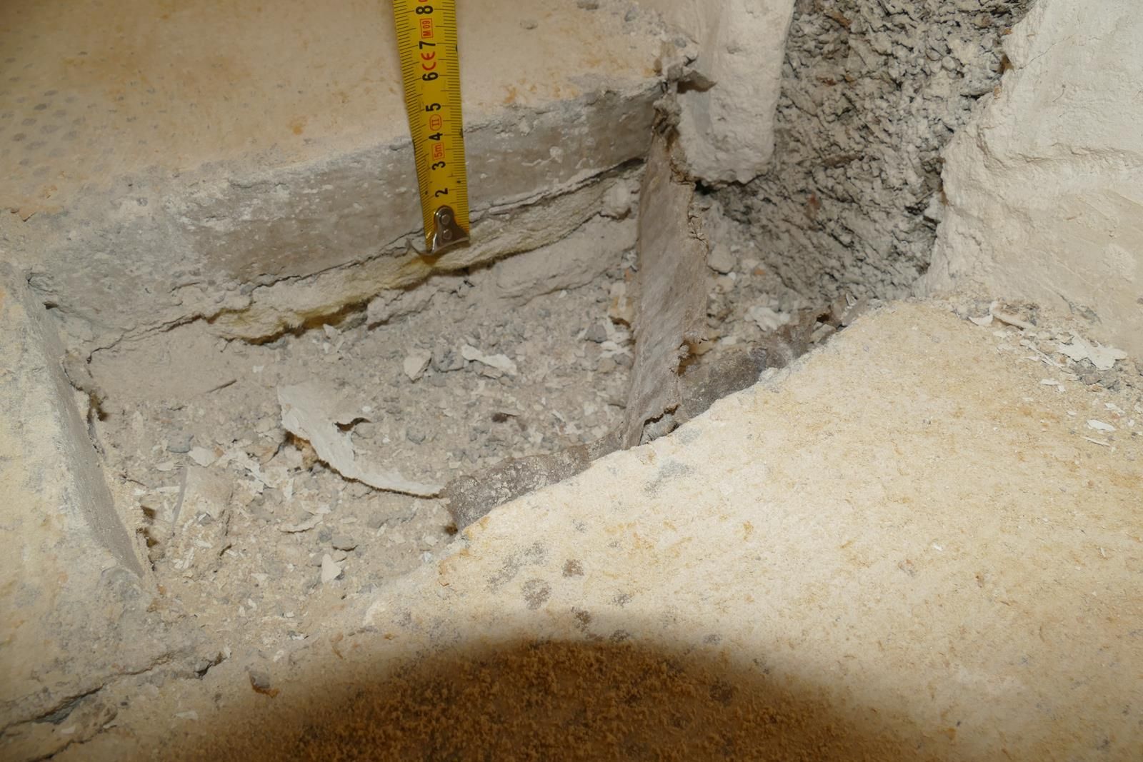 Ein gelbes Maßband, das in ein rechteckiges Loch im Betonboden gesteckt ist, gibt den Blick auf das darunterliegende Material frei.