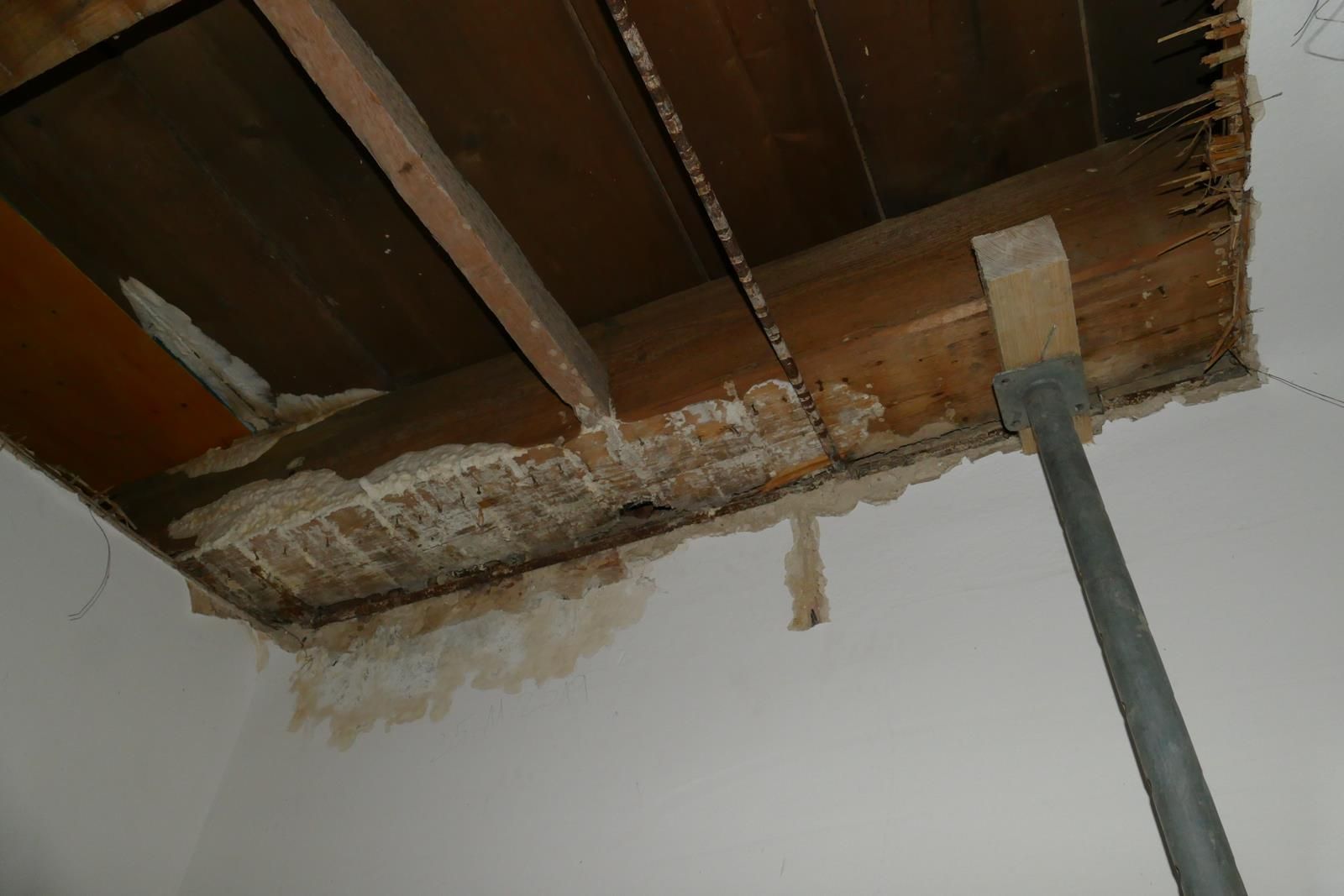 Eine beschädigte Decke, an der freiliegende Holzbalken, Mauerwerk, ein metallener Stützpfosten und Spuren von Wasserflecken zu sehen sind.