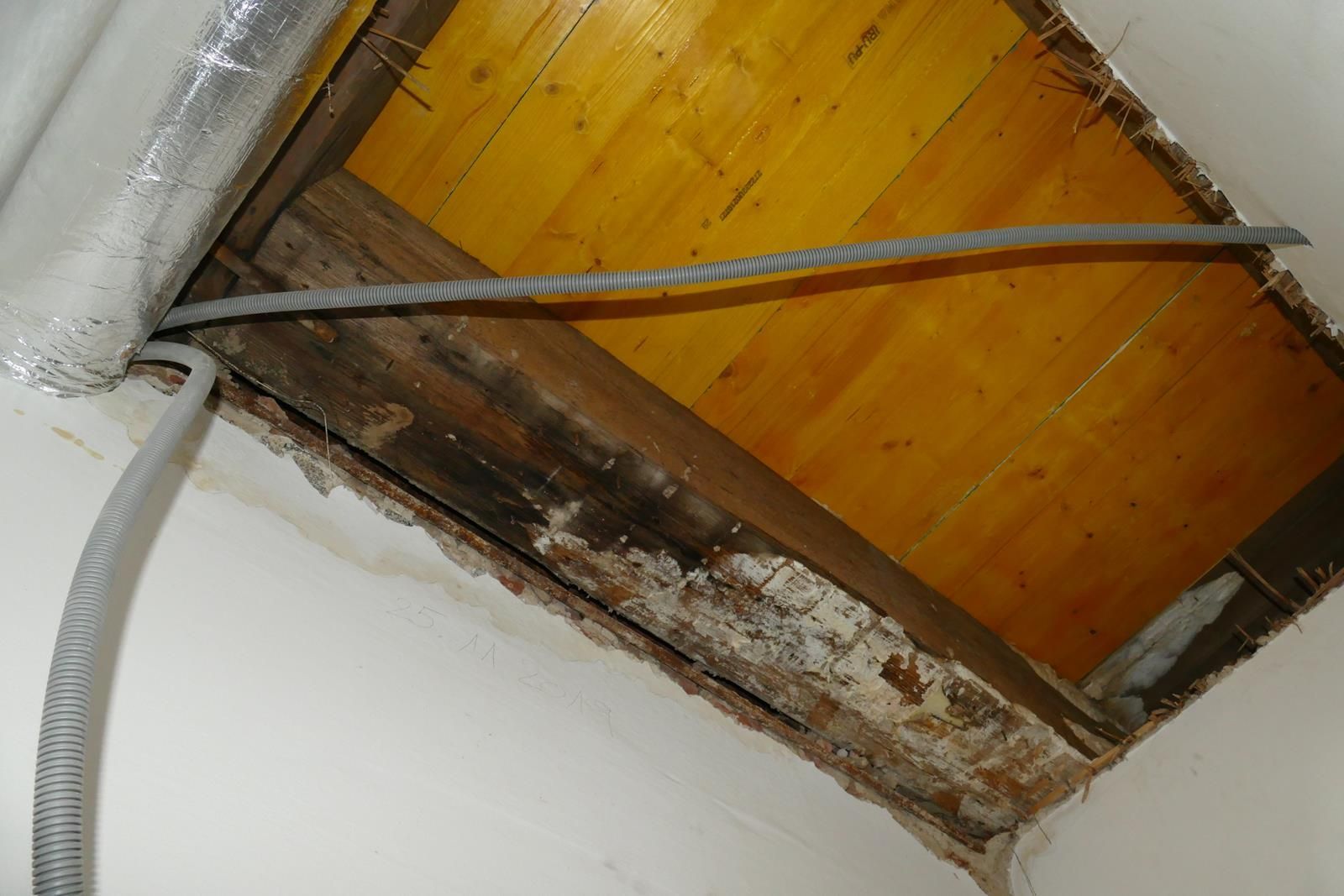 Ein Loch in der Decke gibt den Blick auf Holzbalken, Dielen und ein herabhängendes graues Elektrokabel frei.