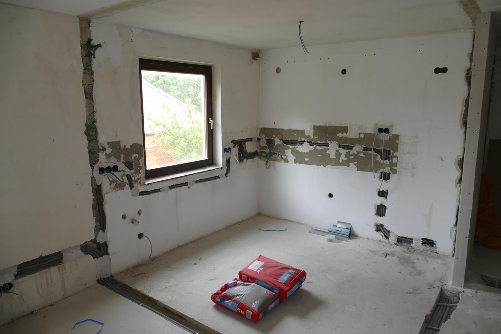 Ein Innenraum im Umbau, mit freiliegenden Kabelkanälen in weißen Wänden, einem Fenster und Baumaterialien.