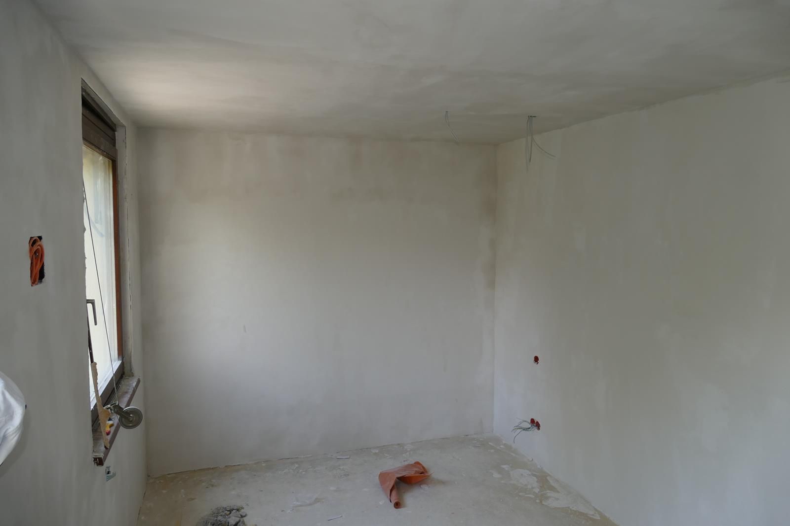 Ein leerer Raum, der gerade renoviert wird, mit weiß verputzten Wänden, einer Decke, einem Fenster und Bauschutt auf dem Boden.