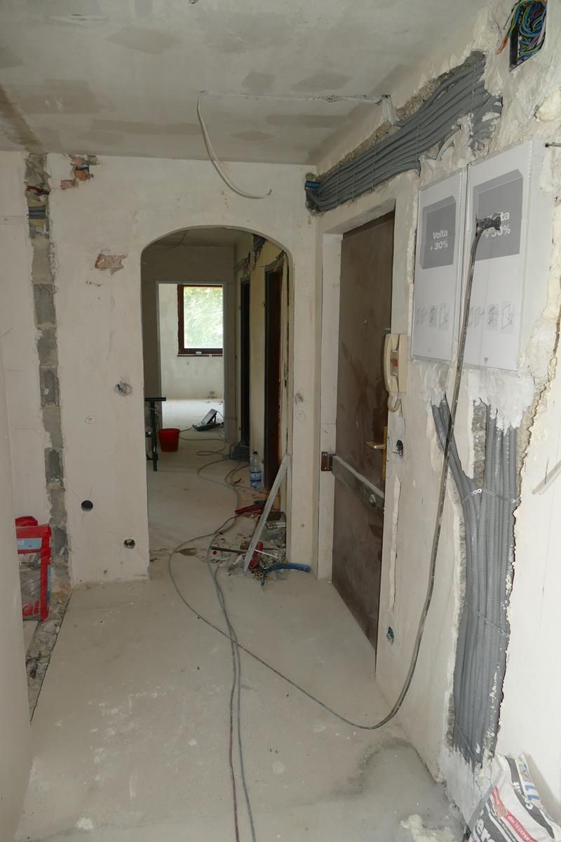 Ein Flur, der gerade renoviert wird, mit freiliegenden elektrischen Leitungen, Mörtelflecken an den Wänden und einem gewölbten Türbogen.