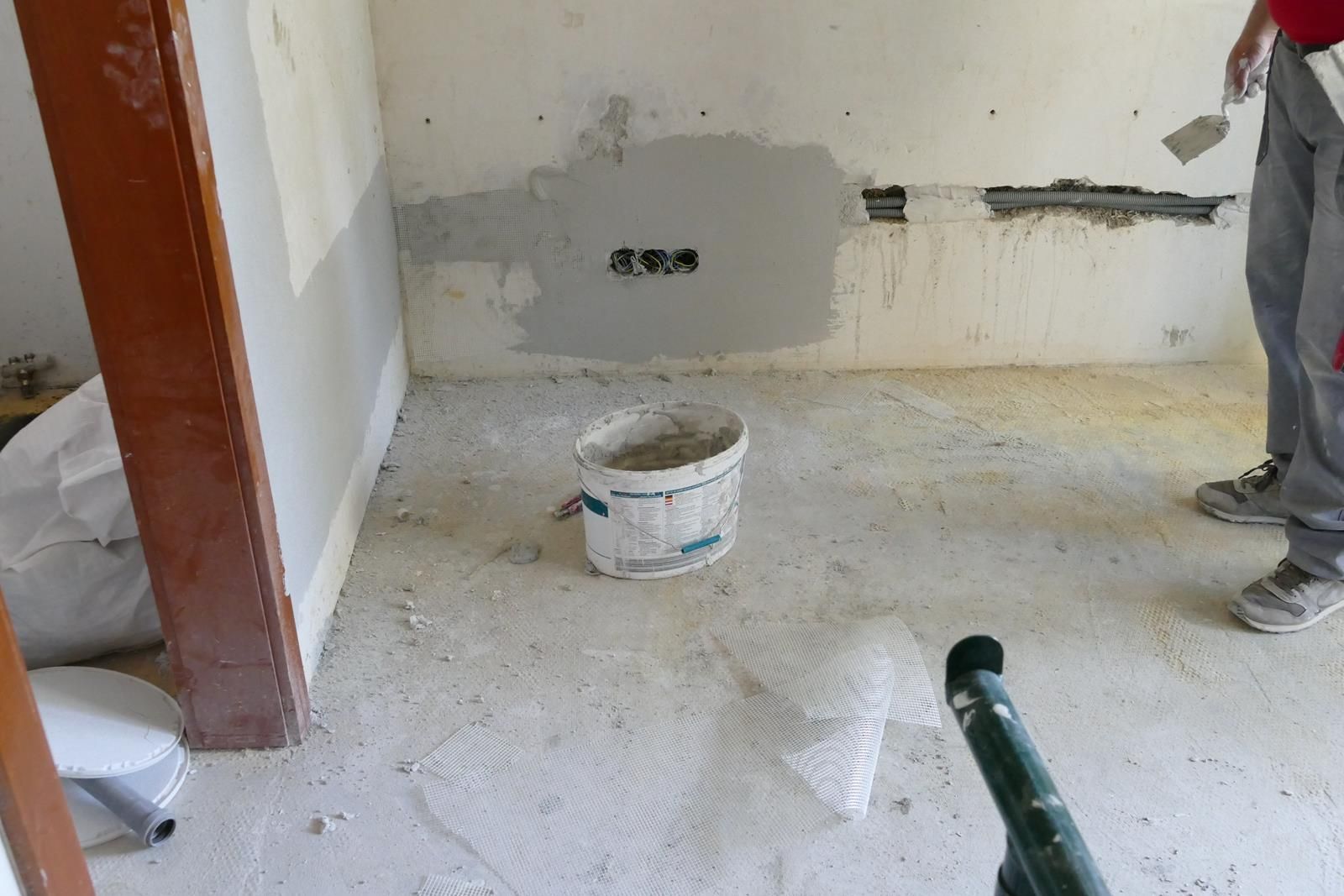 Eine Person mit einer Kelle in der Hand steht in einem Raum, der gerade renoviert wird; neben einer Wand mit freiliegenden Kabeln steht ein Eimer Gips.