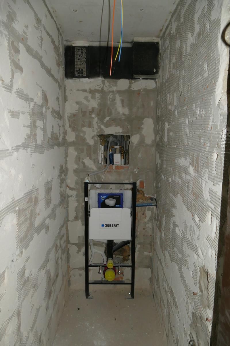 Ein an der Wand montierter Toiletteninstallationsrahmen wurde in einem schmalen, unfertigen Betonraum mit freiliegenden Kabeln an der Oberseite installiert.