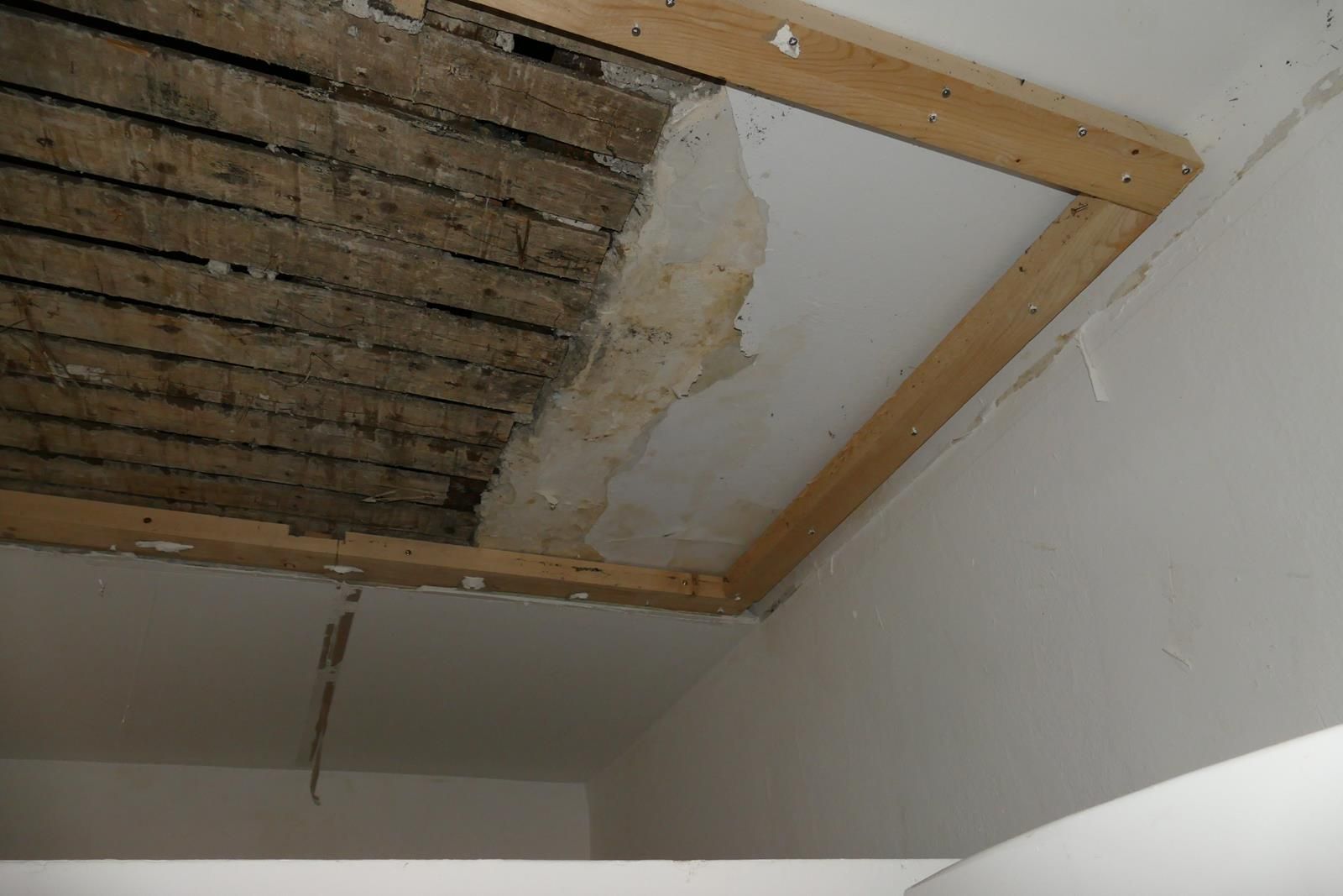 Ein Deckenausschnitt zeigt freiliegende Holzlatten neben einer teilweise ausgebesserten Gipskartonwand, die von Holzbalken eingefasst wird.