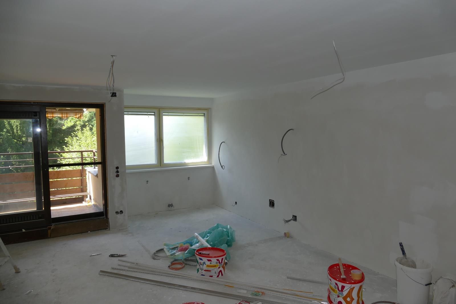 Innenansicht eines im Bau befindlichen Zimmers mit frisch verputzten weißen Wänden, freiliegenden Kabeln und Malutensilien.